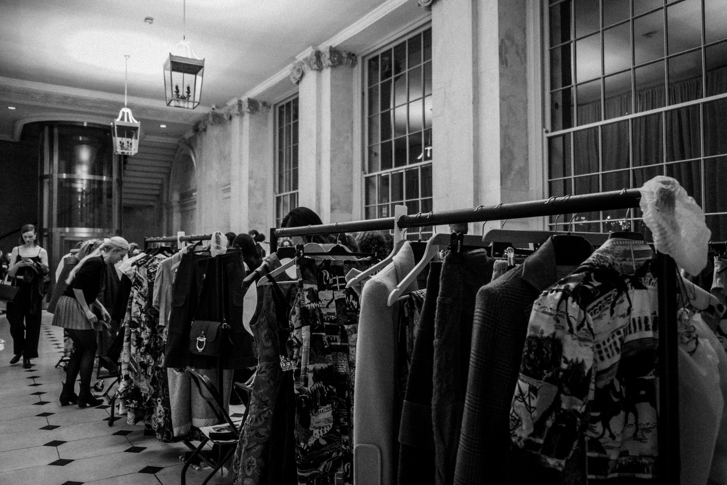 People browsing clothing on racks at an indoor event or sale in a well-lit, elegant room with high ceilings and large windows.