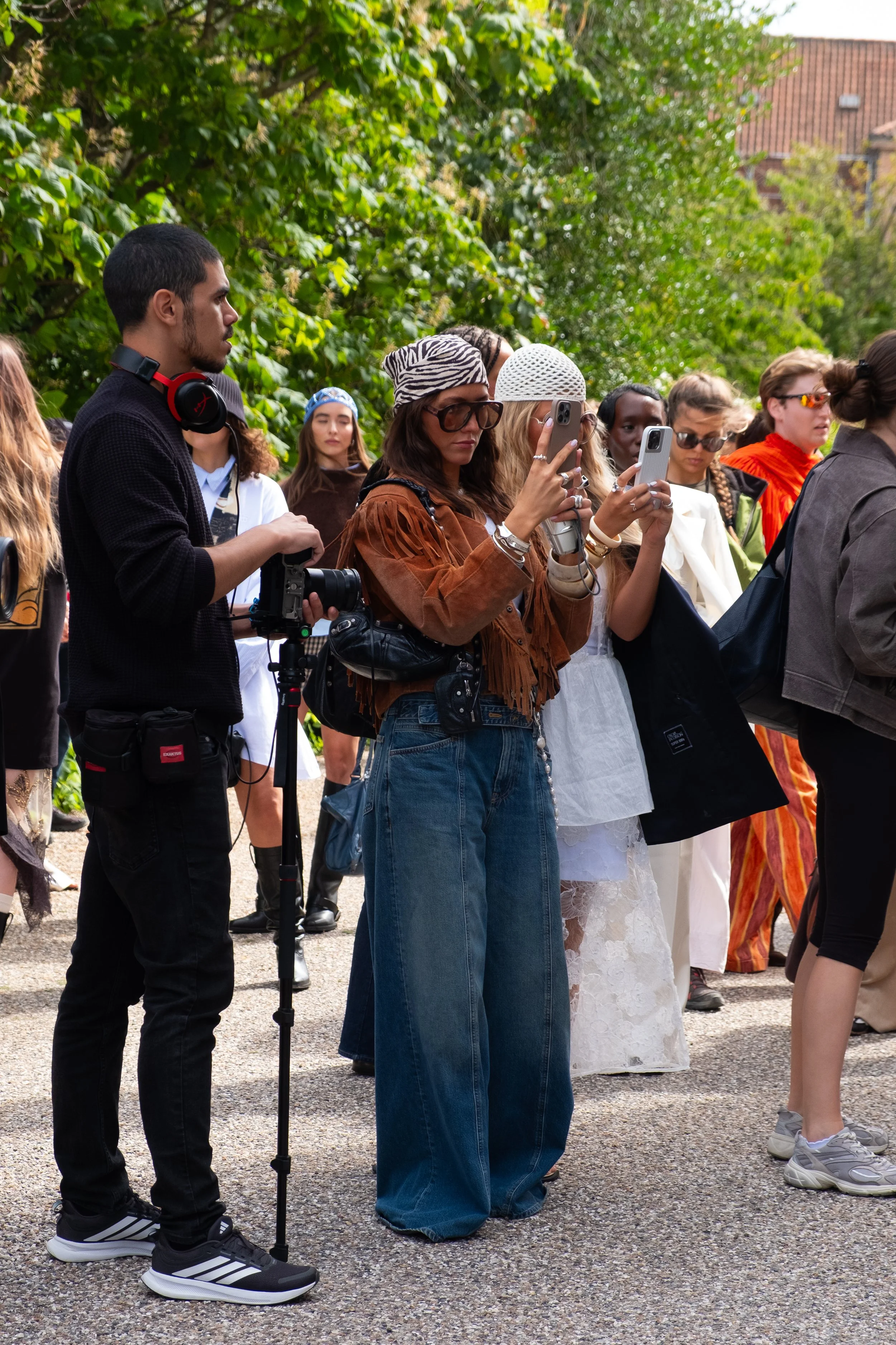 Group of people standing outdoors, some taking photos or videos with smartphones, others observing, with greenery and a brick building in the background.