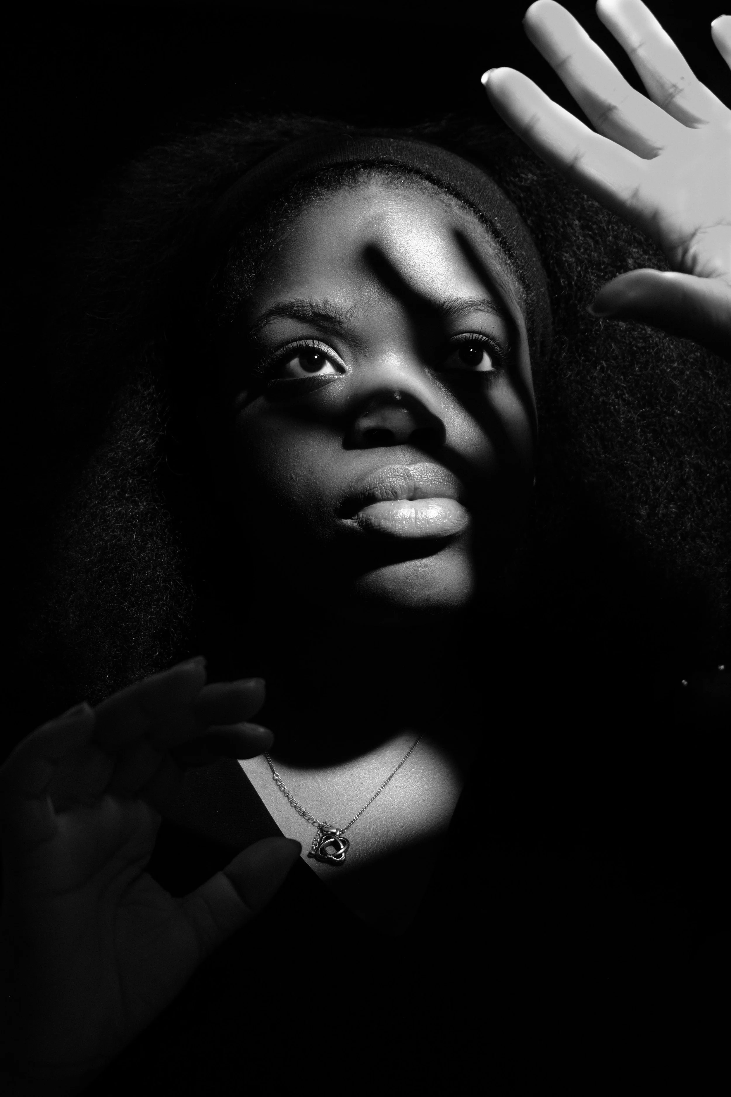 Black and white portrait of a woman with natural hair, wearing a necklace, with shadows cast across her face and hands.