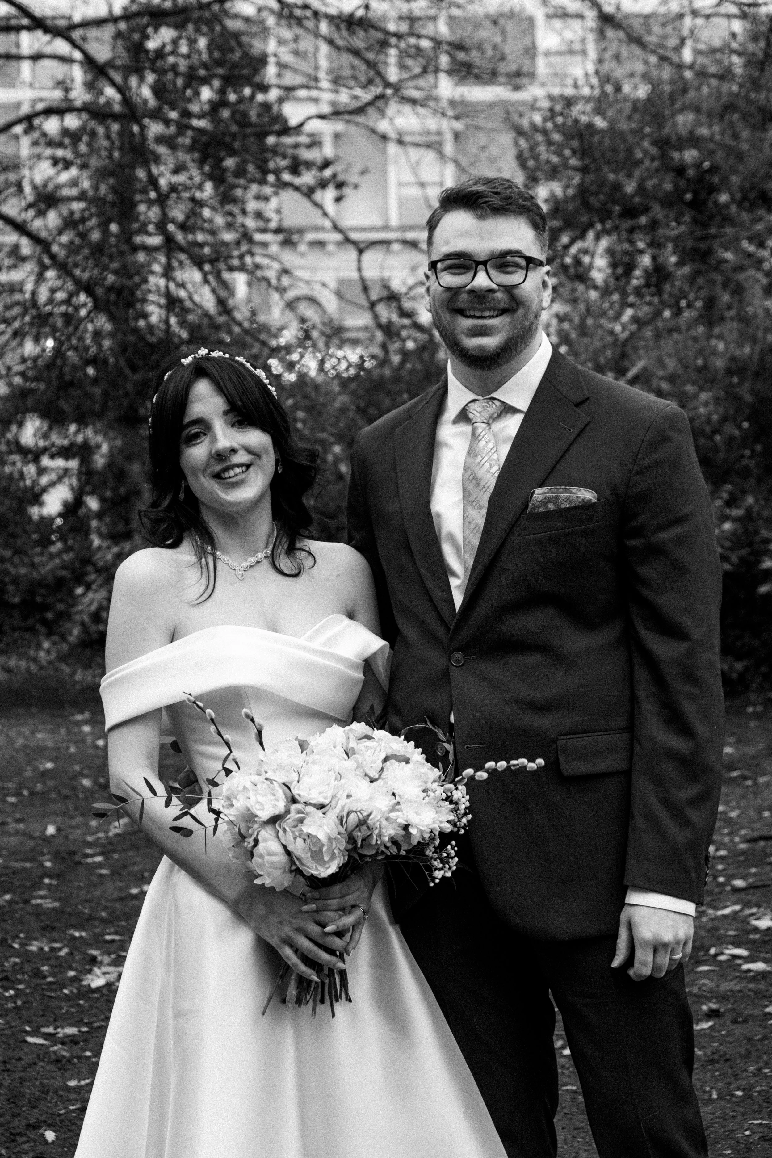 A black-and-white photo of a bride and groom smiling outdoors, with the bride holding a bouquet of flowers and wearing an off-the-shoulder wedding dress, while the groom is dressed in a suit with a tie and pocket square, set against a background of t