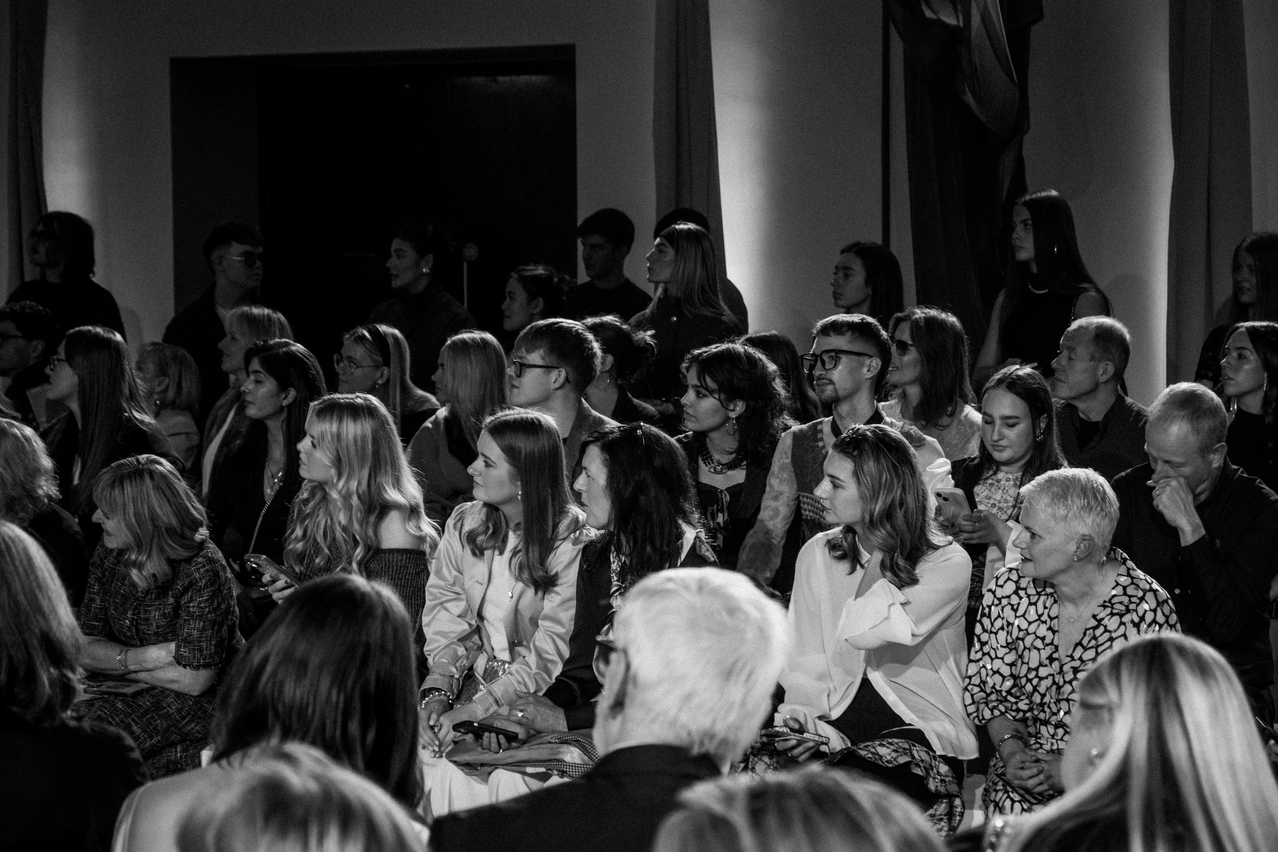 A black and white photo of a diverse group of people sitting and standing, attentively listening during an indoor event or presentation.