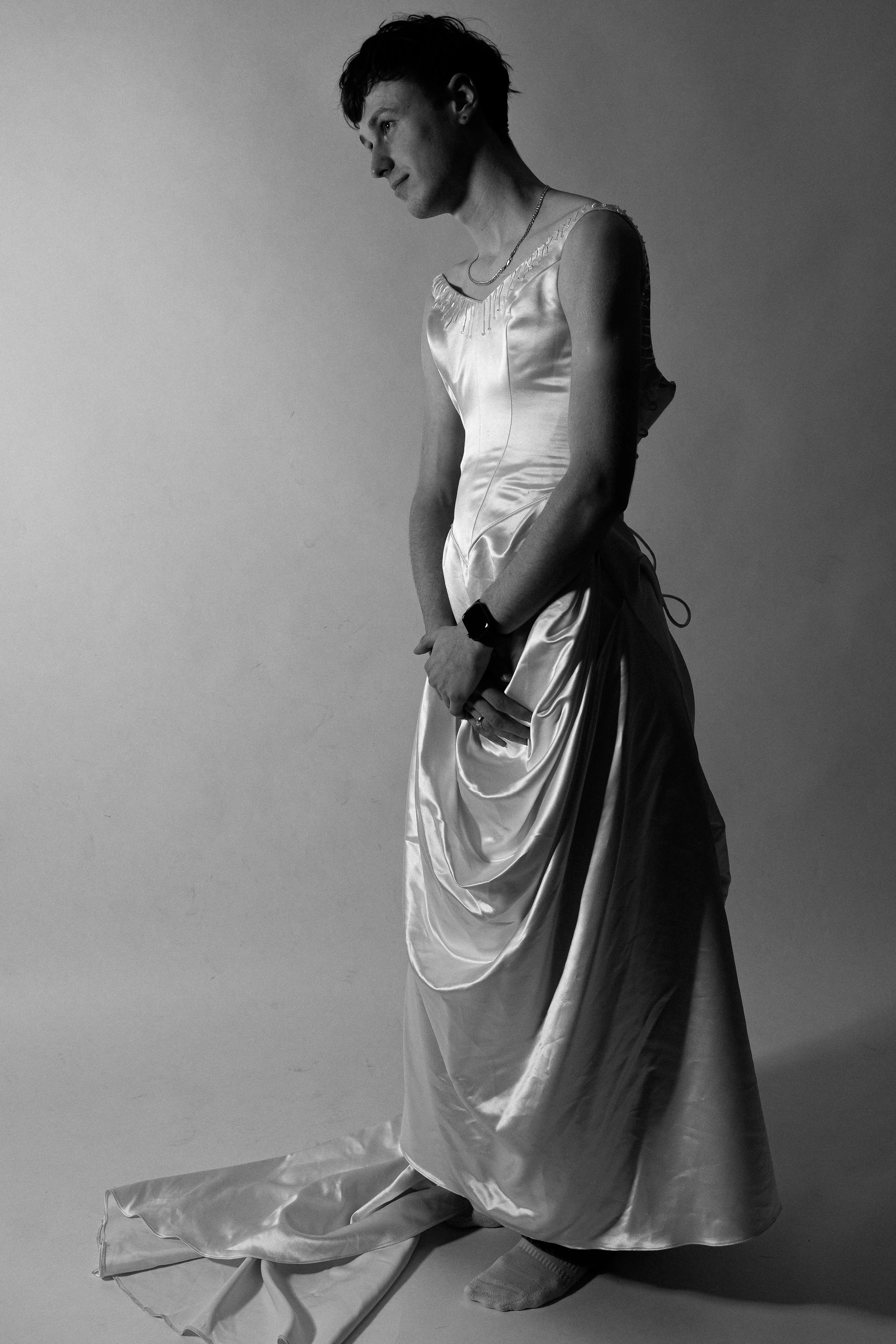 Black and white photo of a person in a satin gown, standing sideways, with a contemplative expression. They are wearing a necklace and a smartwatch, with the gown's train on the floor.
