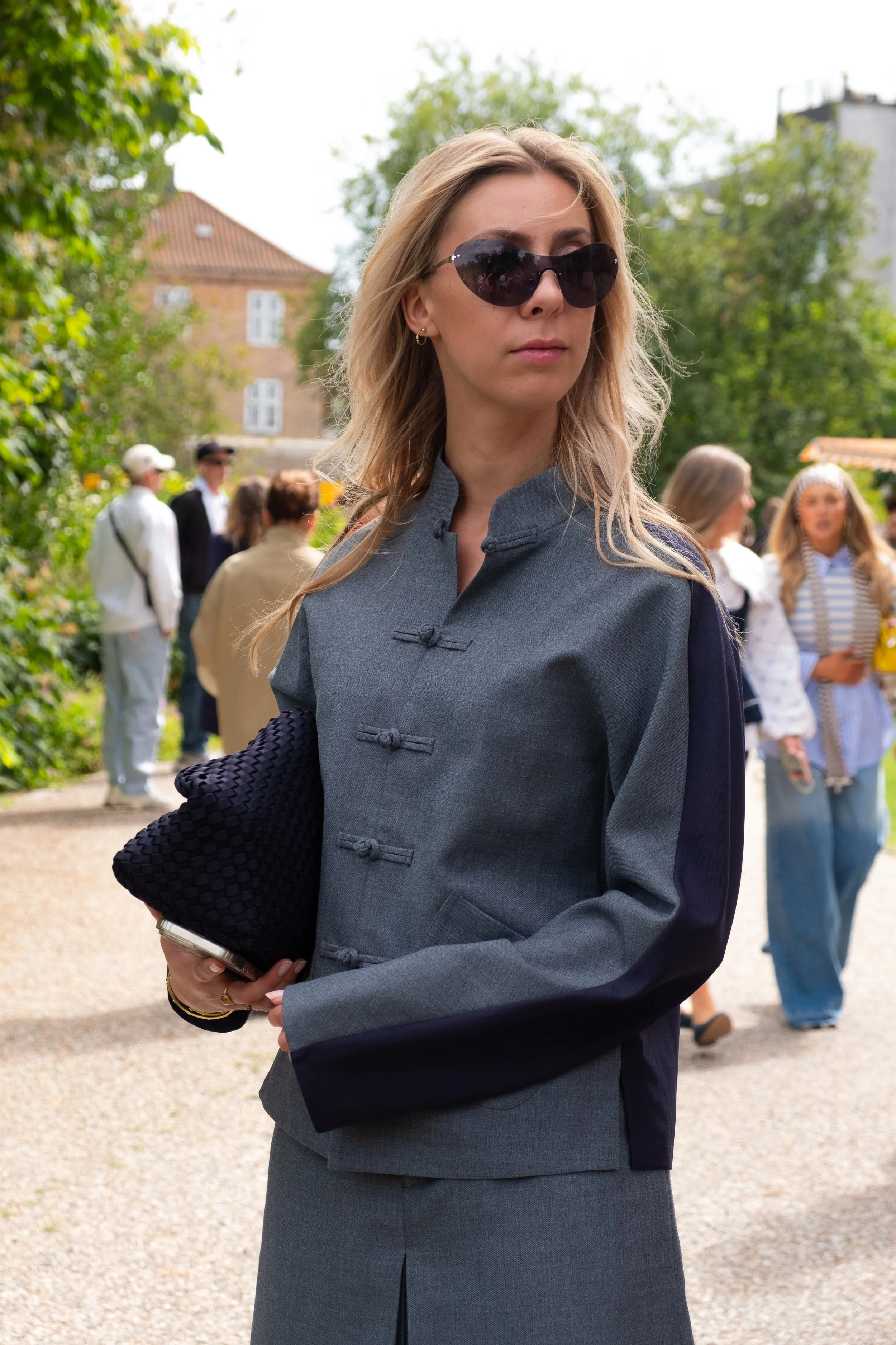 A woman wearing dark sunglasses and a traditional-style gray jacket with knotted buttons stands outdoors among trees and a group of people.