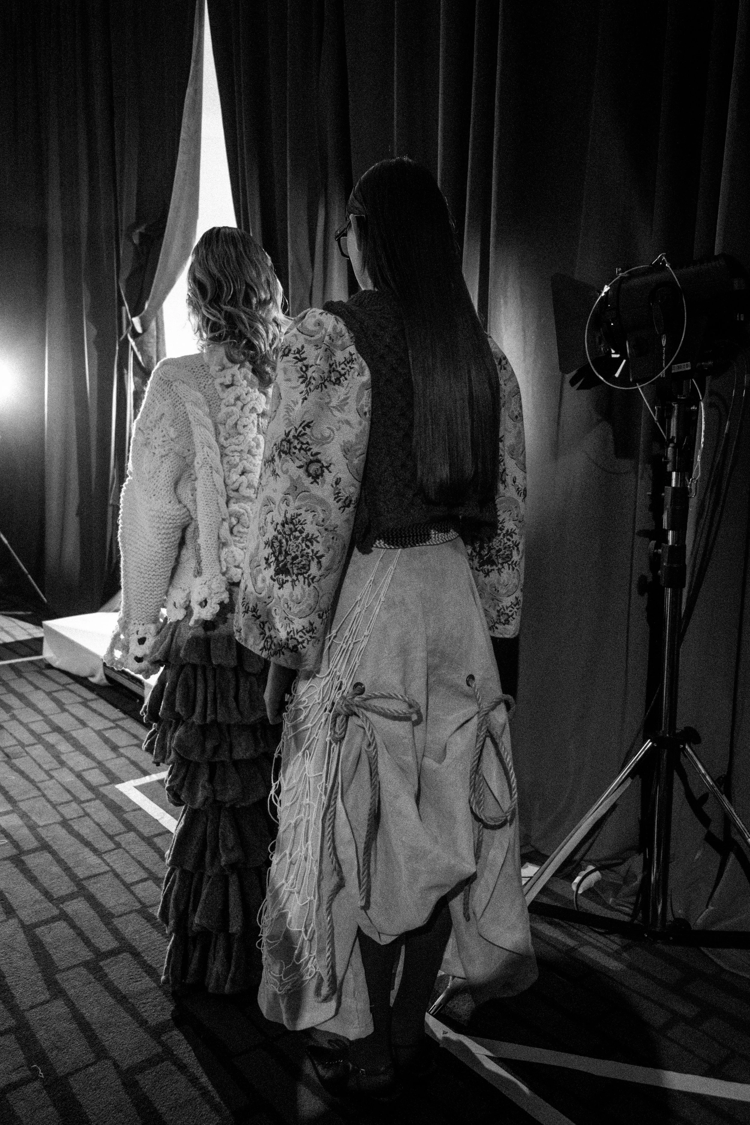 Two women standing behind curtains at a photo shoot or film set, with studio lighting equipment nearby.