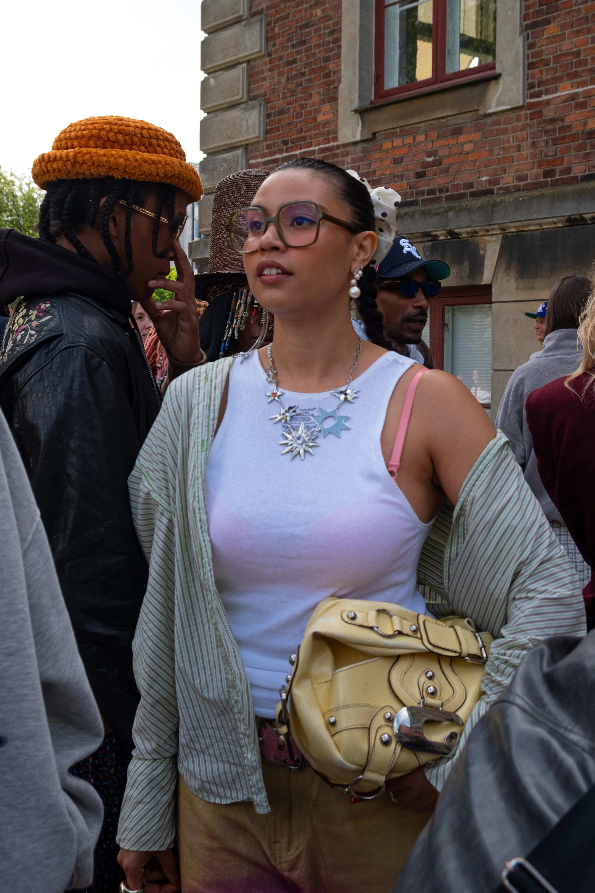 A woman wearing glasses and a white tank top with a necklace, holding a yellow purse, standing outdoors among a crowd of people in front of a brick building.