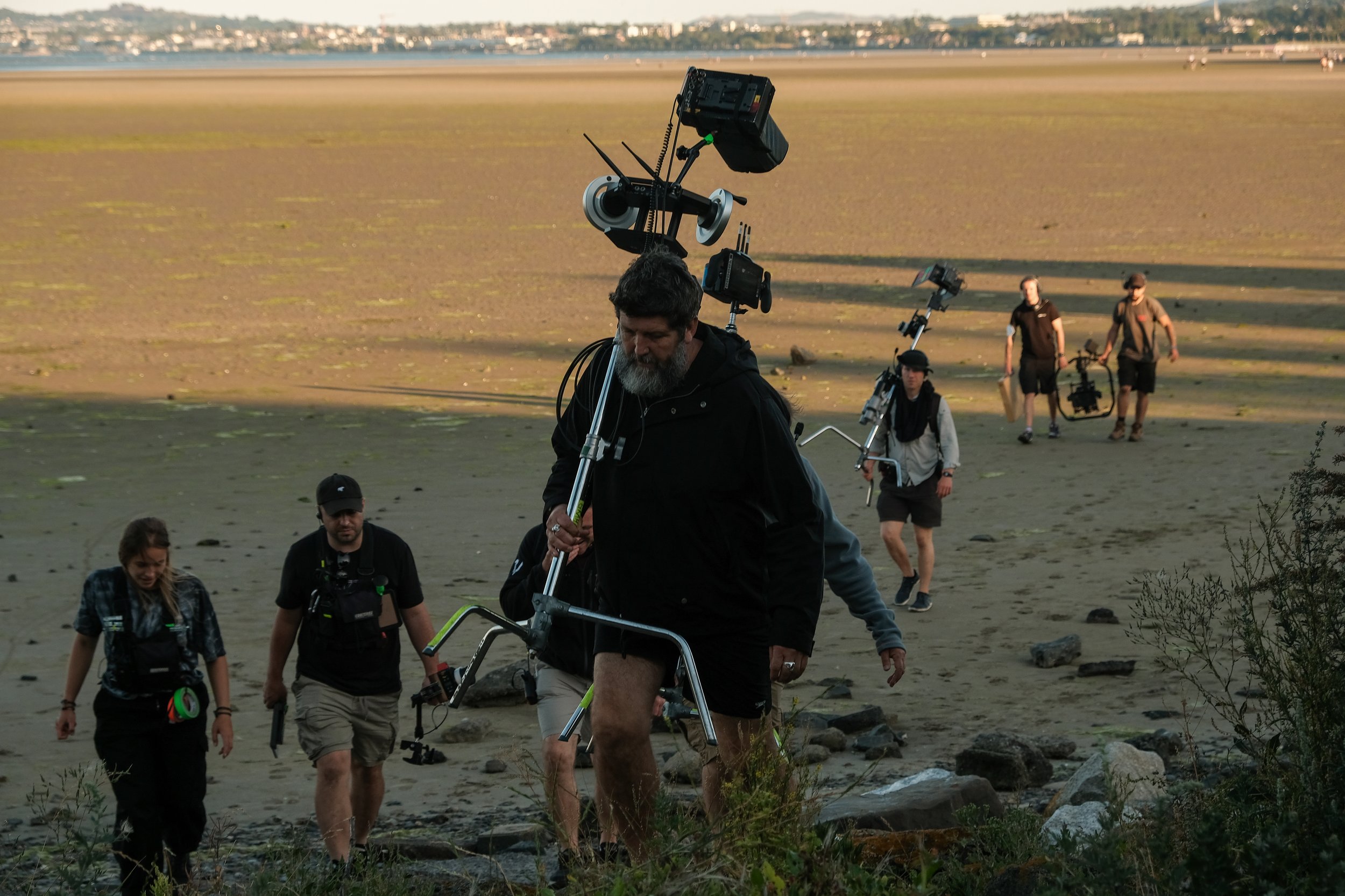 A film crew with cameras and equipment walking across a sandy beach during sunset or sunrise, with a distant city skyline in the background.