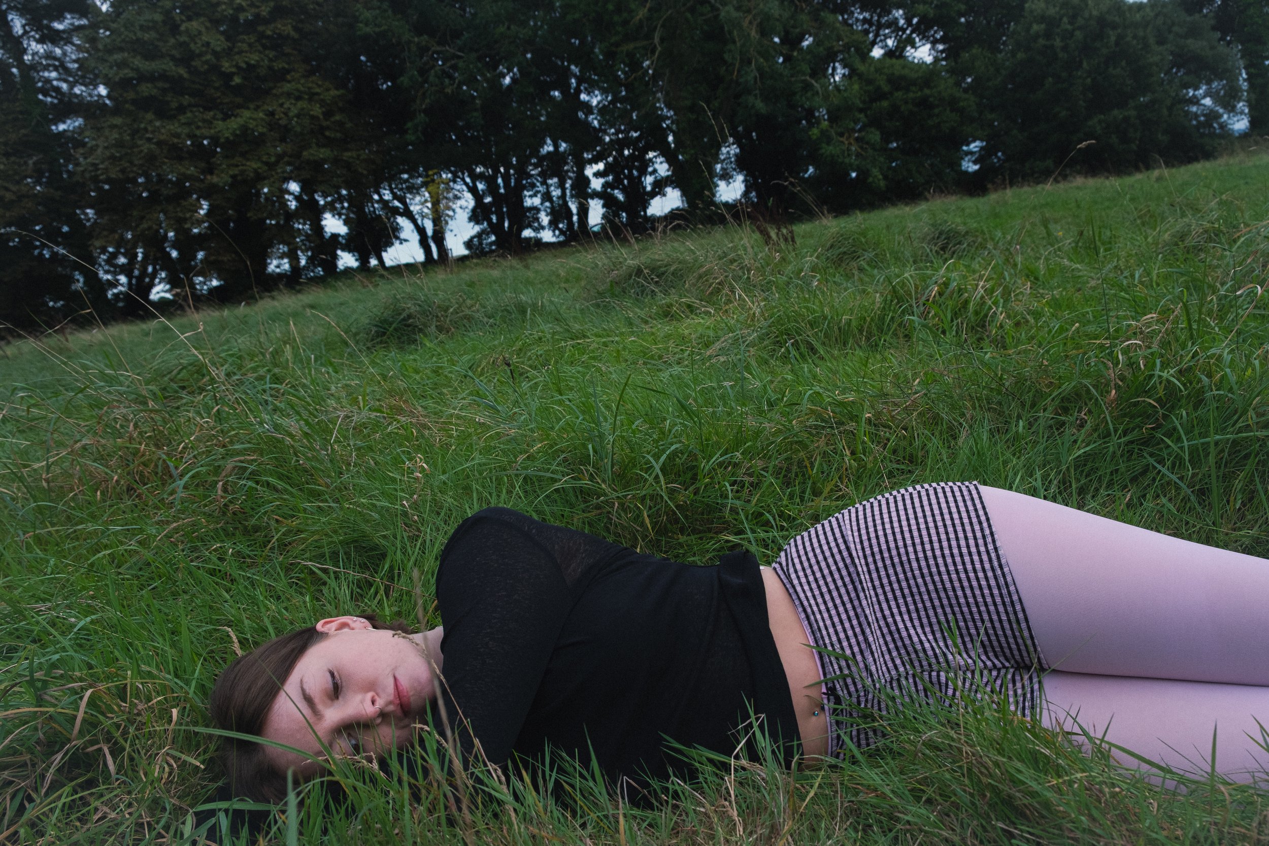 A young woman lies on the grass in a field, eyes closed, with trees in the background.