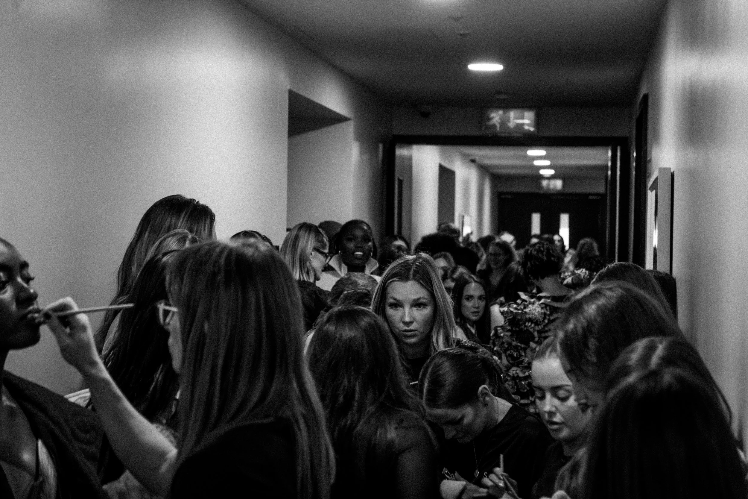 Crowded hallway with many women waiting and socializing, some using phones, in a black and white photograph.