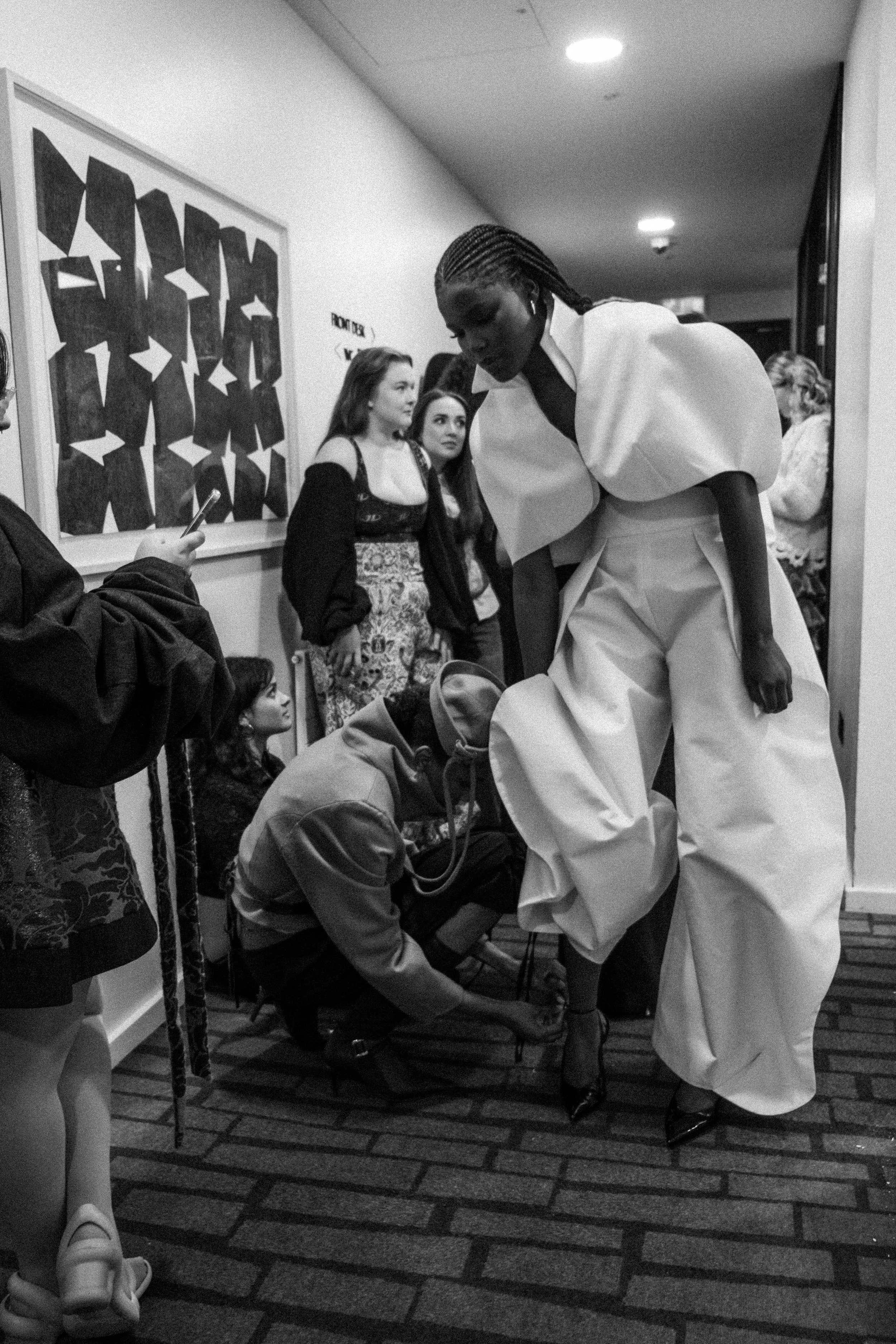 A woman in high fashion clothing adjusting her shoes as she stands among a group of young people in a hallway, with others observing and taking photos.