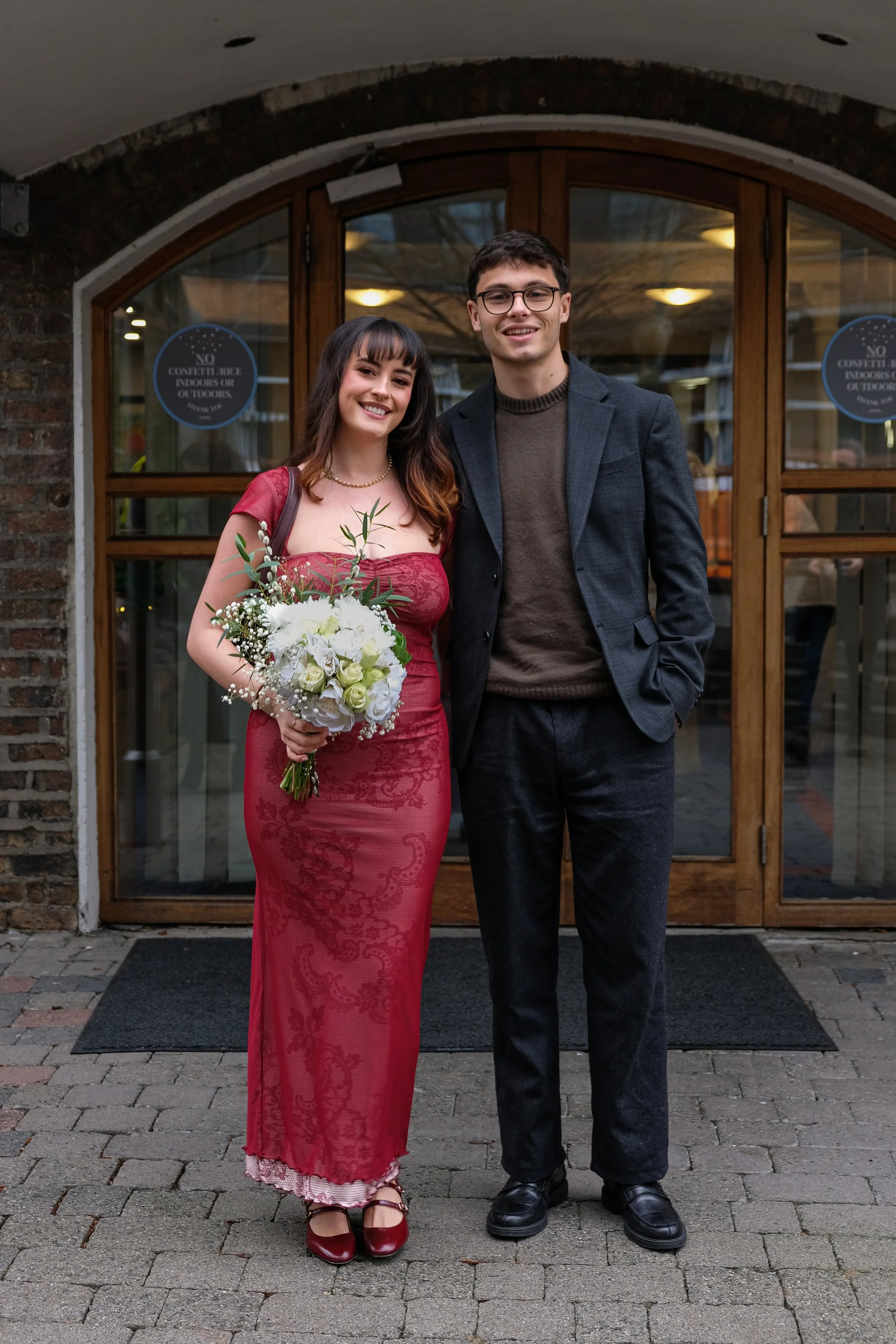 A young couple standing outside a building, the woman wearing a red dress holding a bouquet, and the man wearing a dark blazer and trousers. Both are smiling.