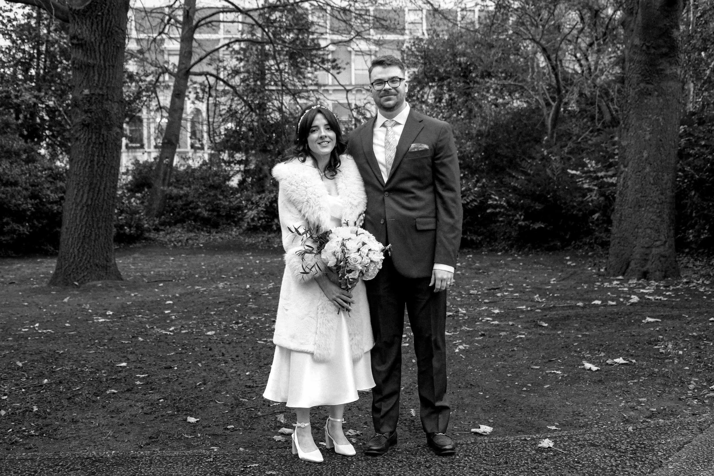Black and white photo of a bride and groom standing outdoors in a park, surrounded by trees, with buildings in the background. The bride holds a bouquet and wears a dress with a fur coat, while the groom wears a suit and tie.