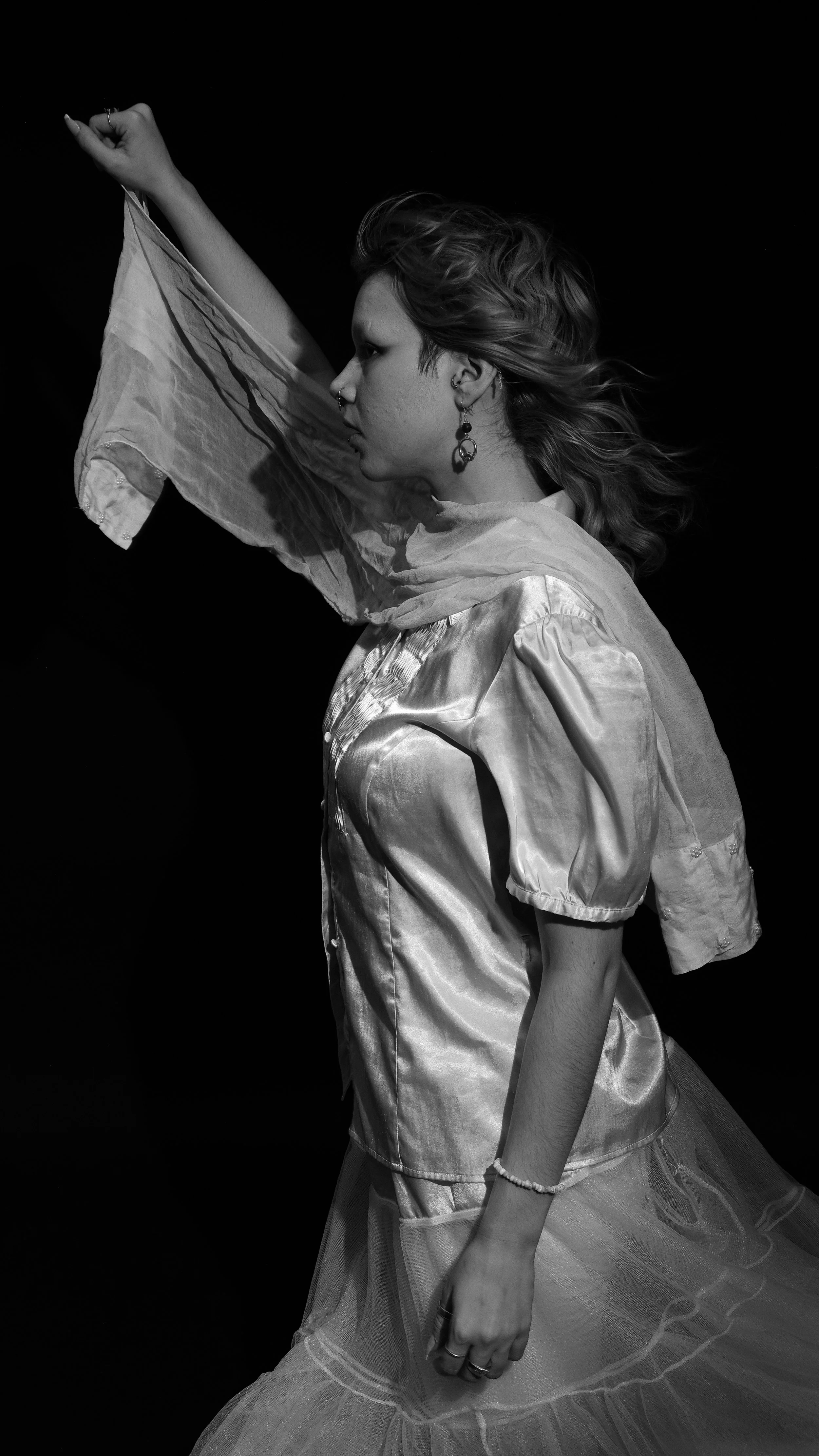 A black-and-white photograph of a woman in vintage clothing, posing with her arm raised and her hair blowing to the side.