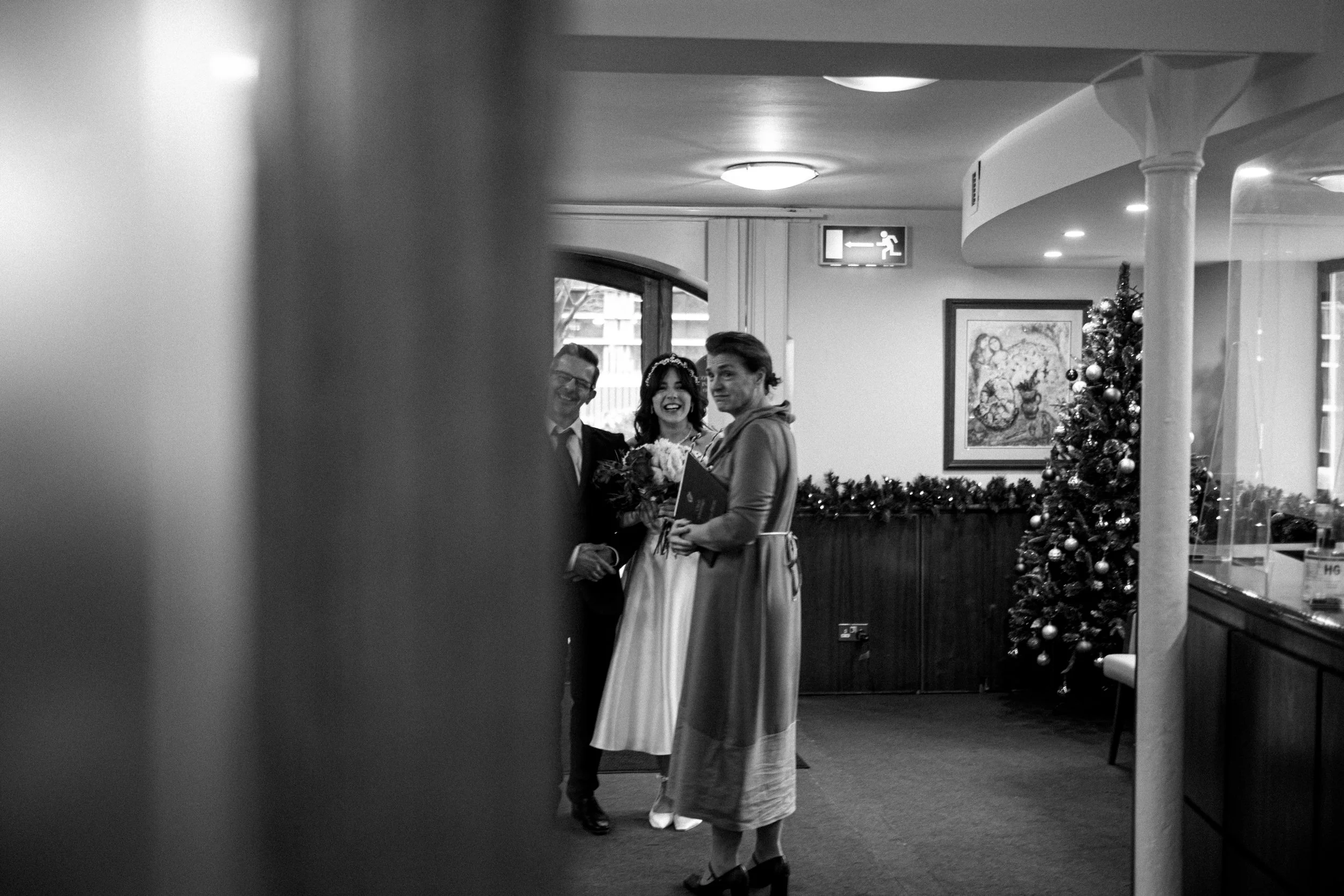 A wedding scene in black and white, featuring a bride with a bouquet, a man in suit, and a woman in a dress, standing near a decorated Christmas tree in a cozy indoor setting.
