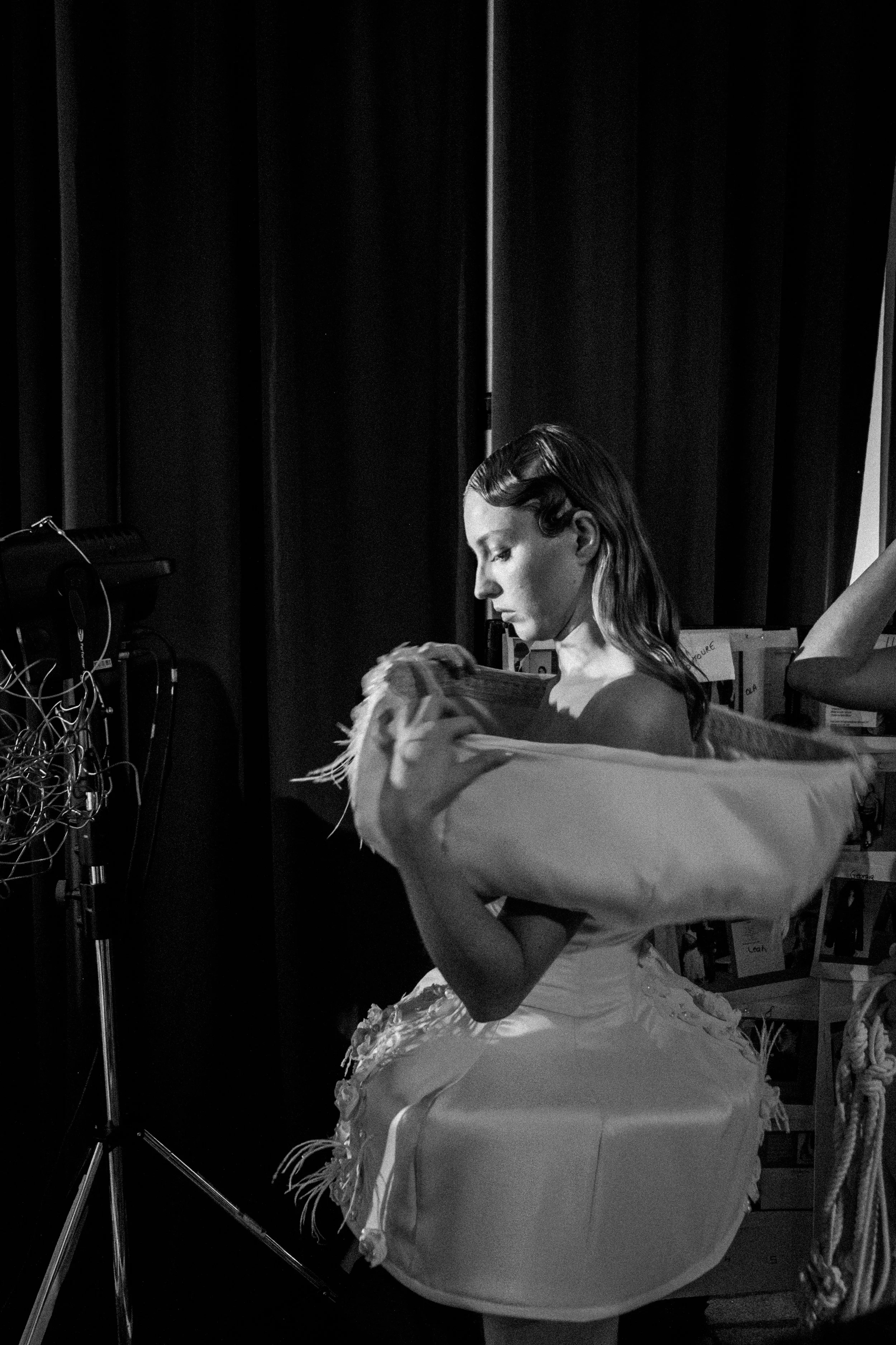 A woman with styled hair and a white dress is preparing for a photoshoot or performance, adjusting a large, feathered costume or prop, in a studio or backstage setting with equipment and boxes.