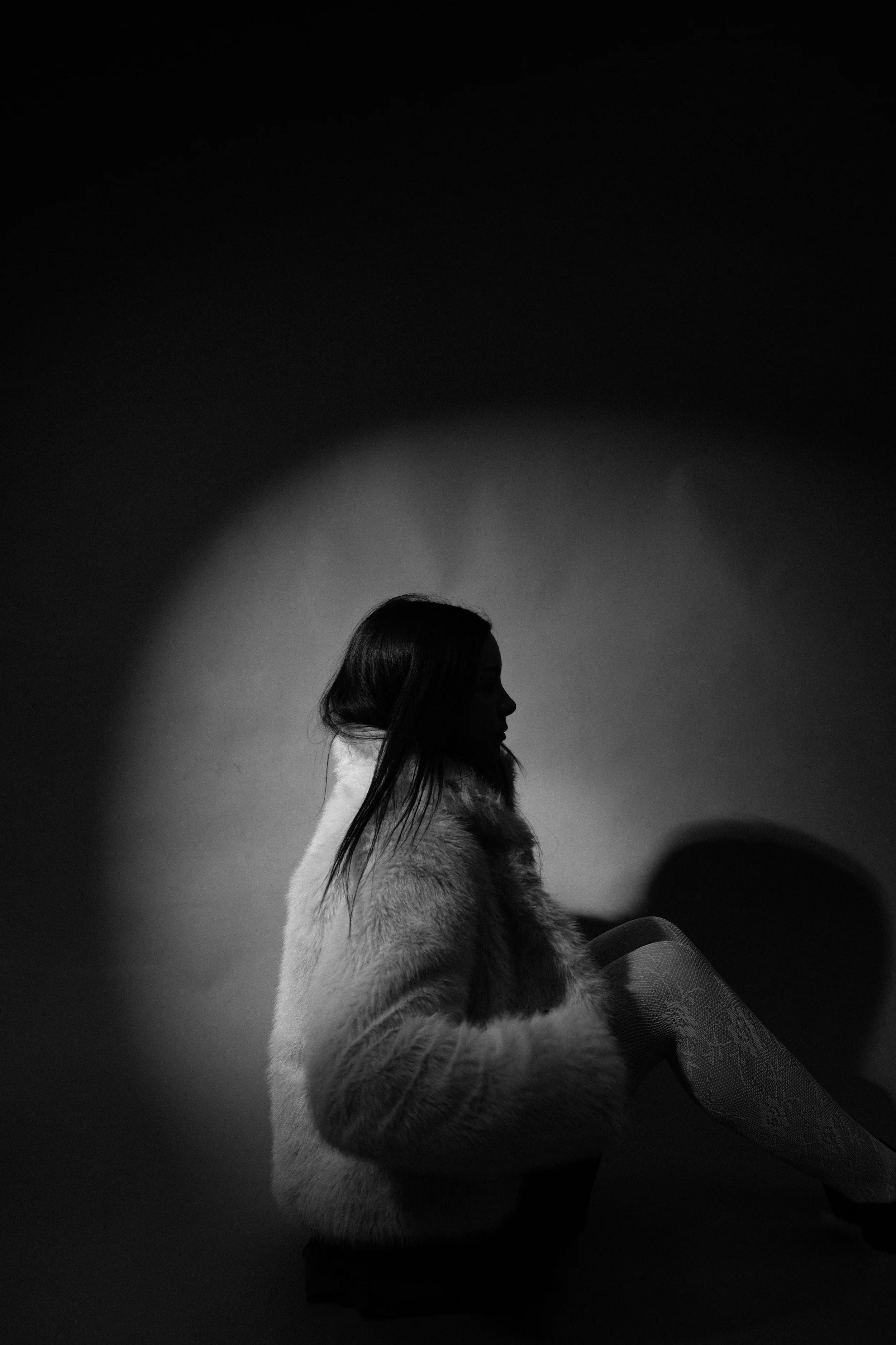 Silhouette of a woman with long hair sitting against a wall, illuminated by a circular light, black and white photo.