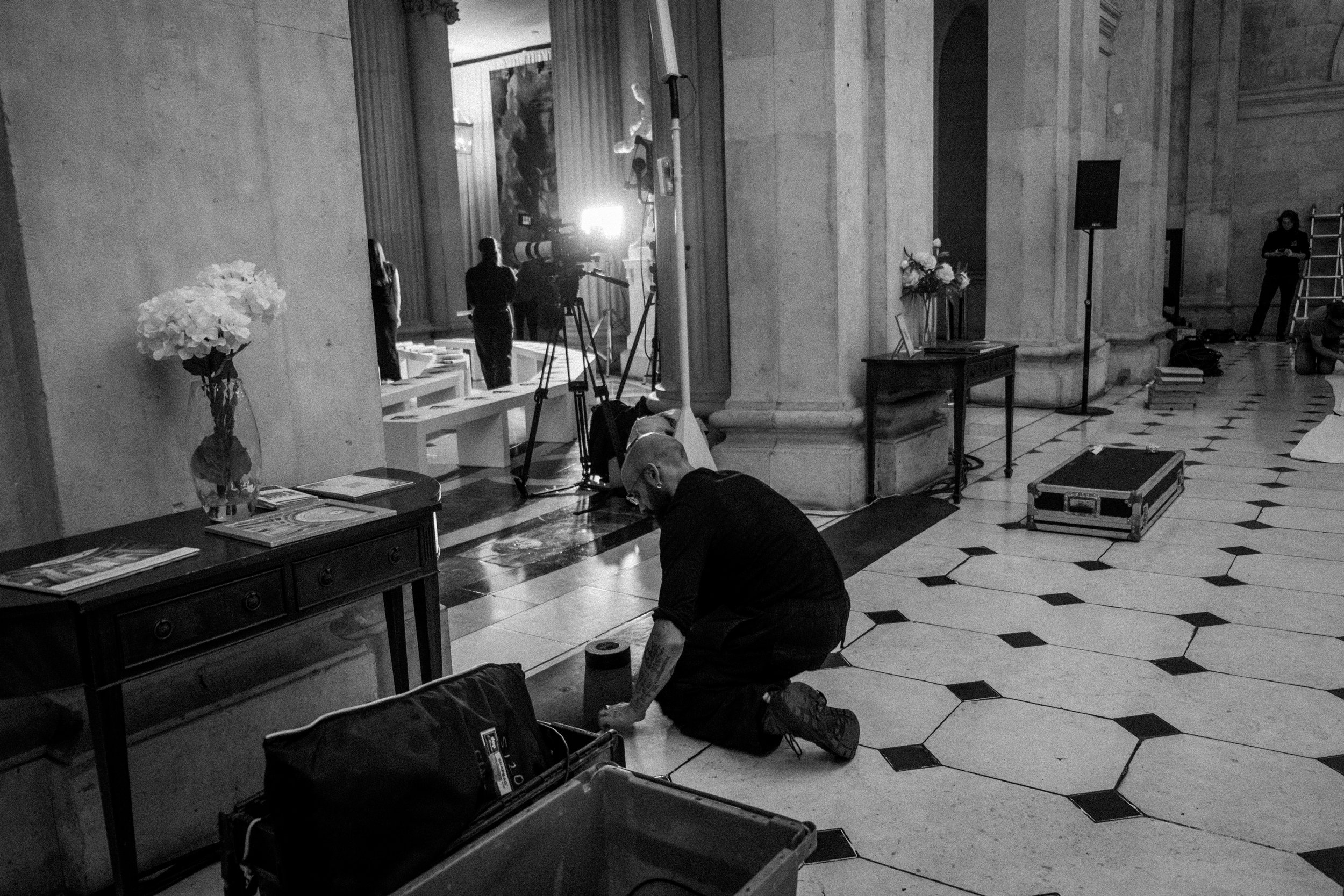 A man kneeling on the floor working with tools in a large indoor space with tall columns, a black and white tile floor, and camera equipment in the background.