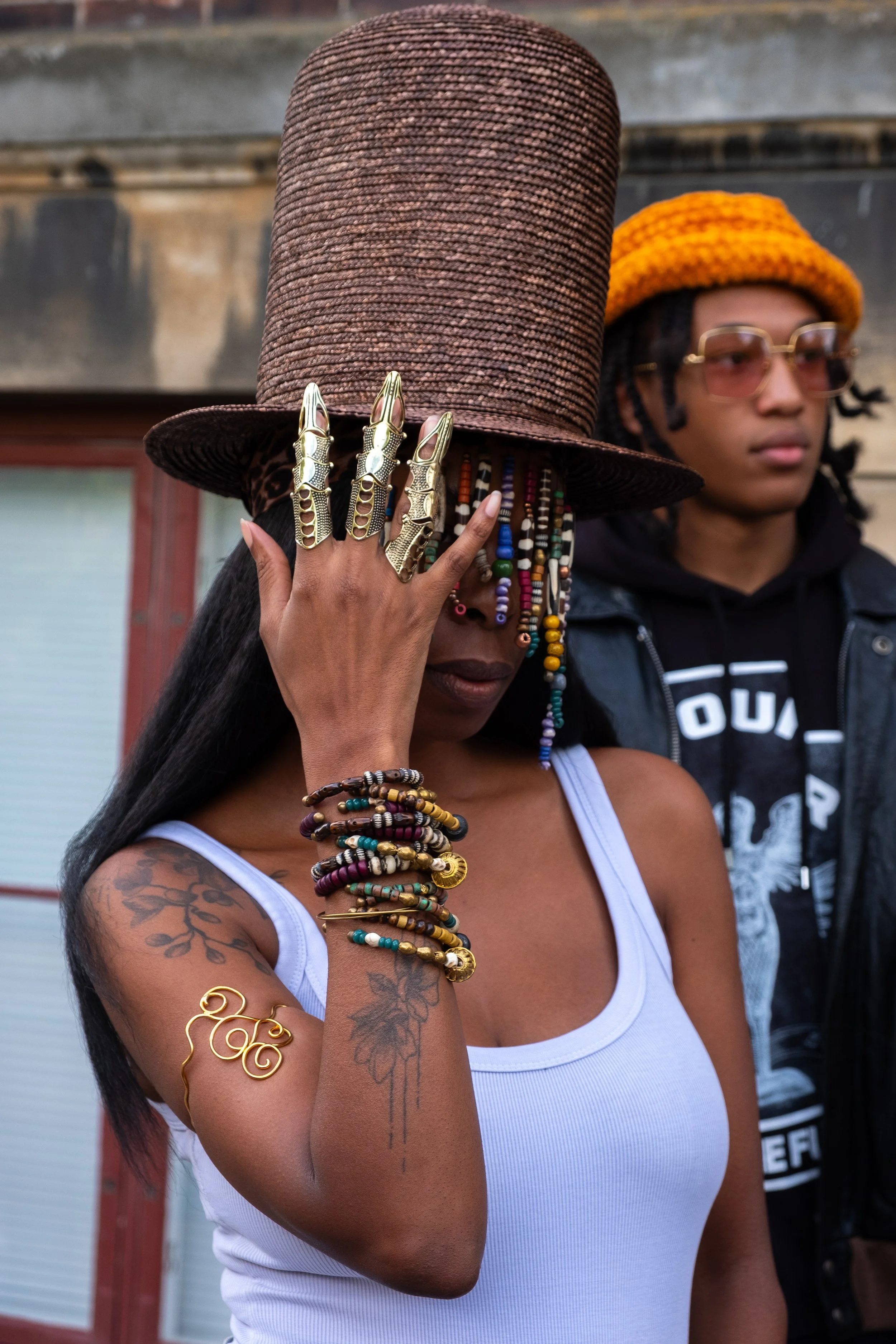A woman wearing a large brown hat, with colorful beaded hair and multiple bracelets, rings, and tattoos, holds her hand over her face. A man with glasses, dreadlocks, and a black jacket stands behind her.