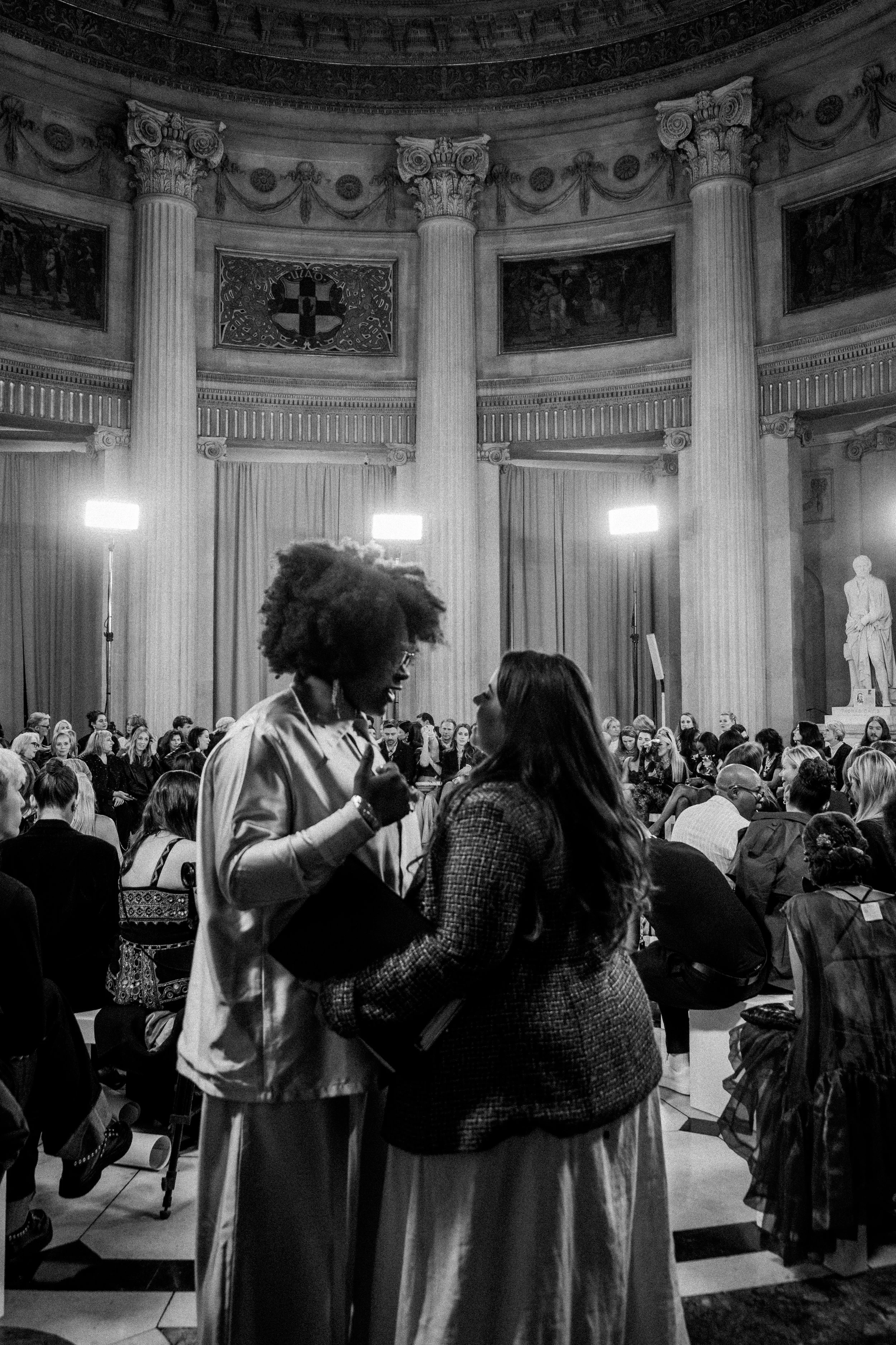 Two women in conversation at an event in a grand hall with tall columns and statues, with many seated guests in the background and bright lights.