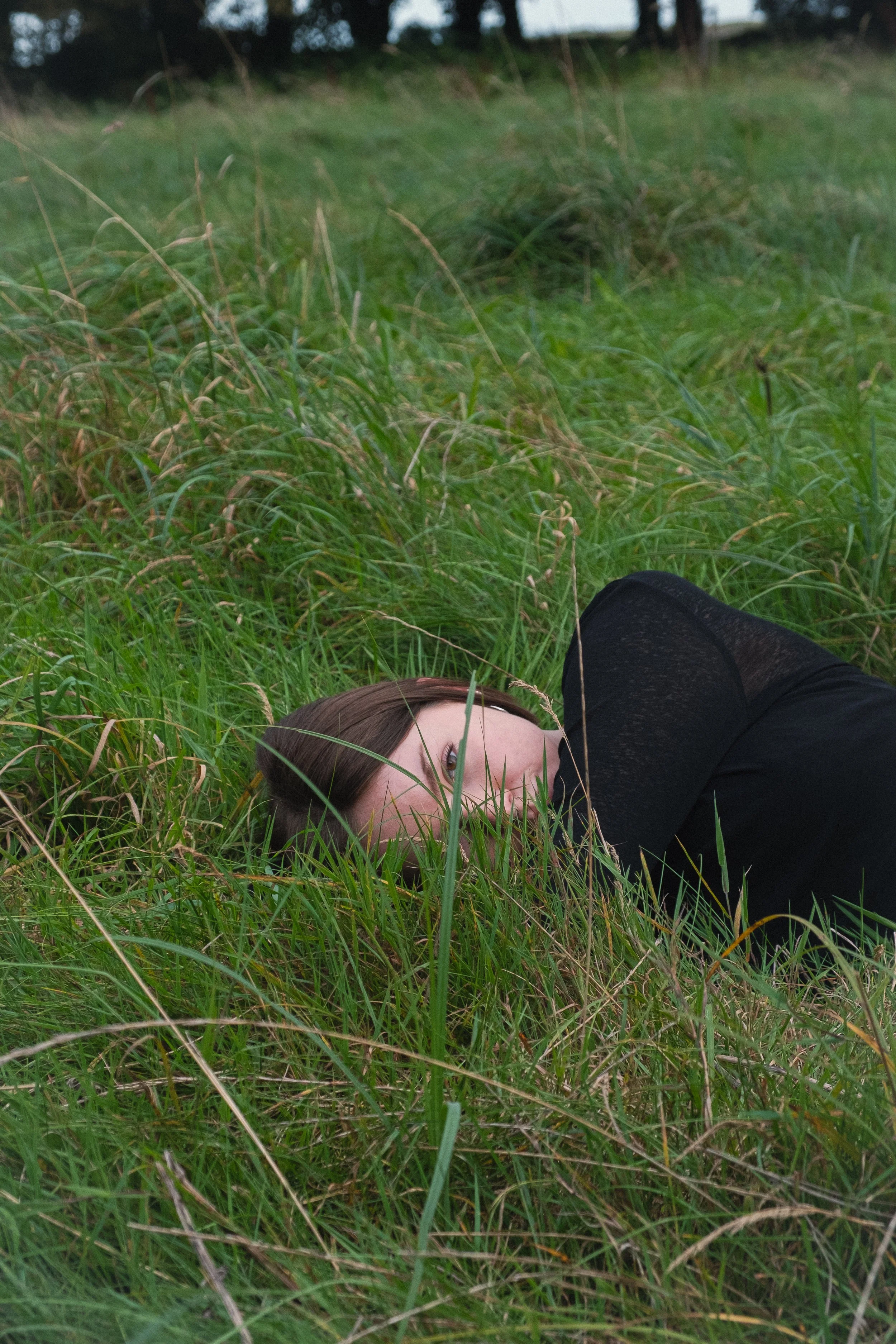 A woman lying on her side in tall green grass with her face partially obscured by blades of grass, wearing a black shirt.
