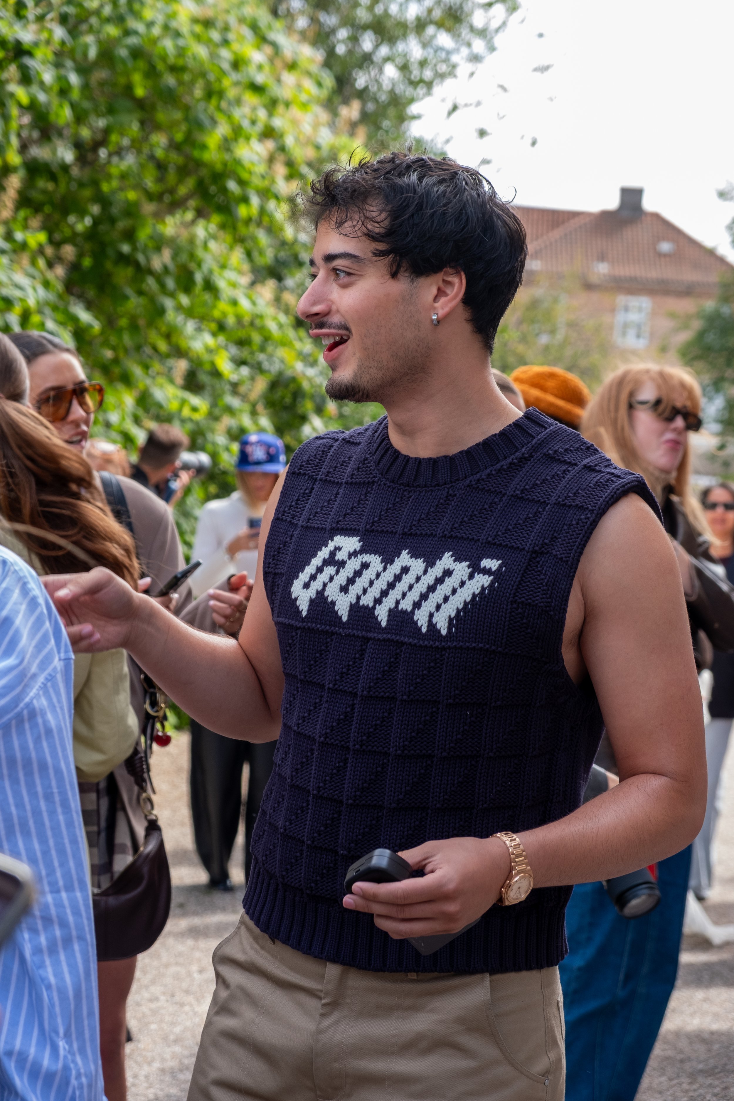Young man with dark hair, wearing a sleeveless navy sweater with white text, holding a phone, engaging with a crowd outdoors.