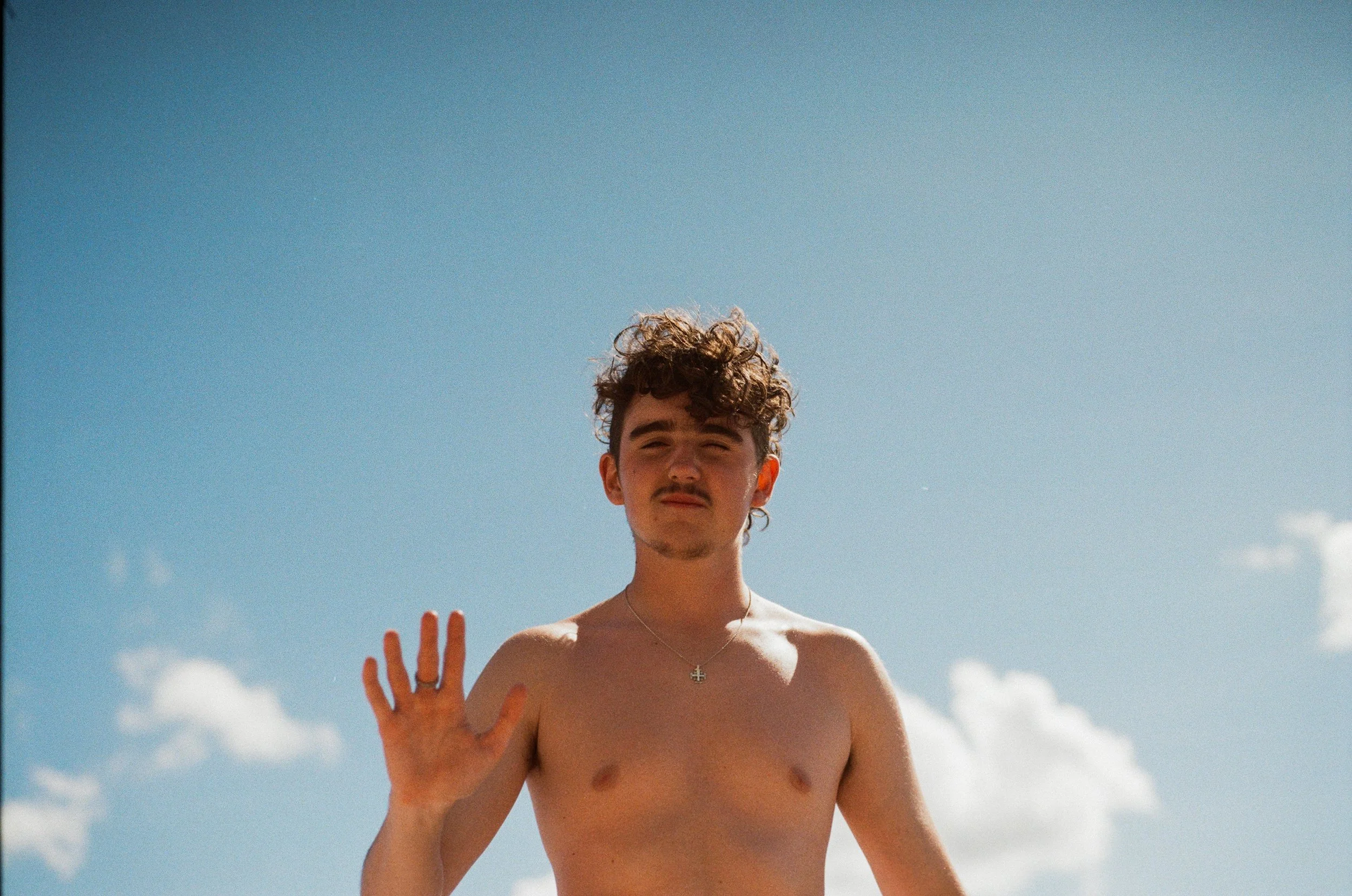 A shirtless young man with curly hair and a cross necklace, standing outdoors under a clear blue sky, waving at the camera.