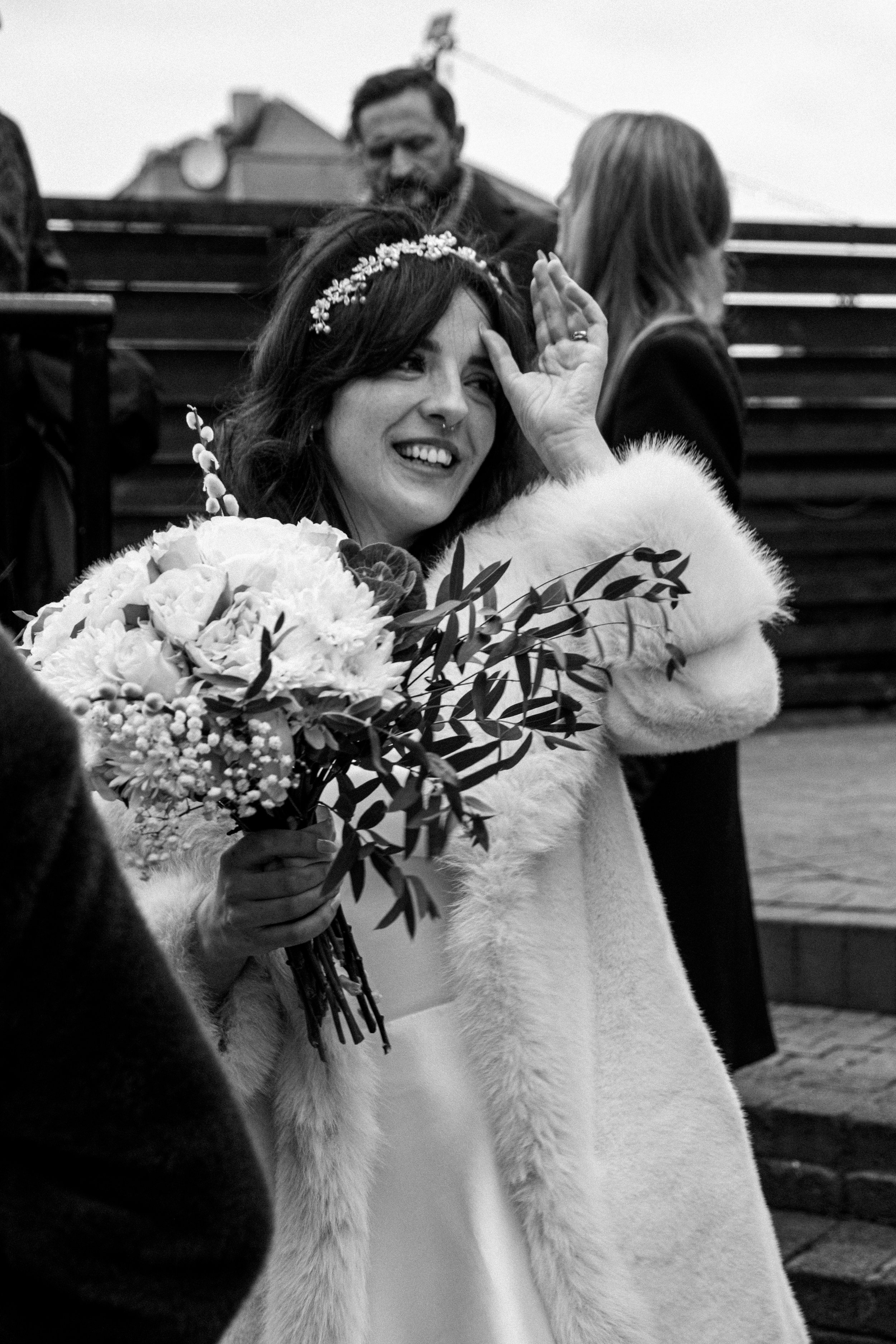 A woman smiling and holding a bouquet of flowers, wearing a headband and a fur coat, is celebrating at an outdoor event, with people and steps in the background.