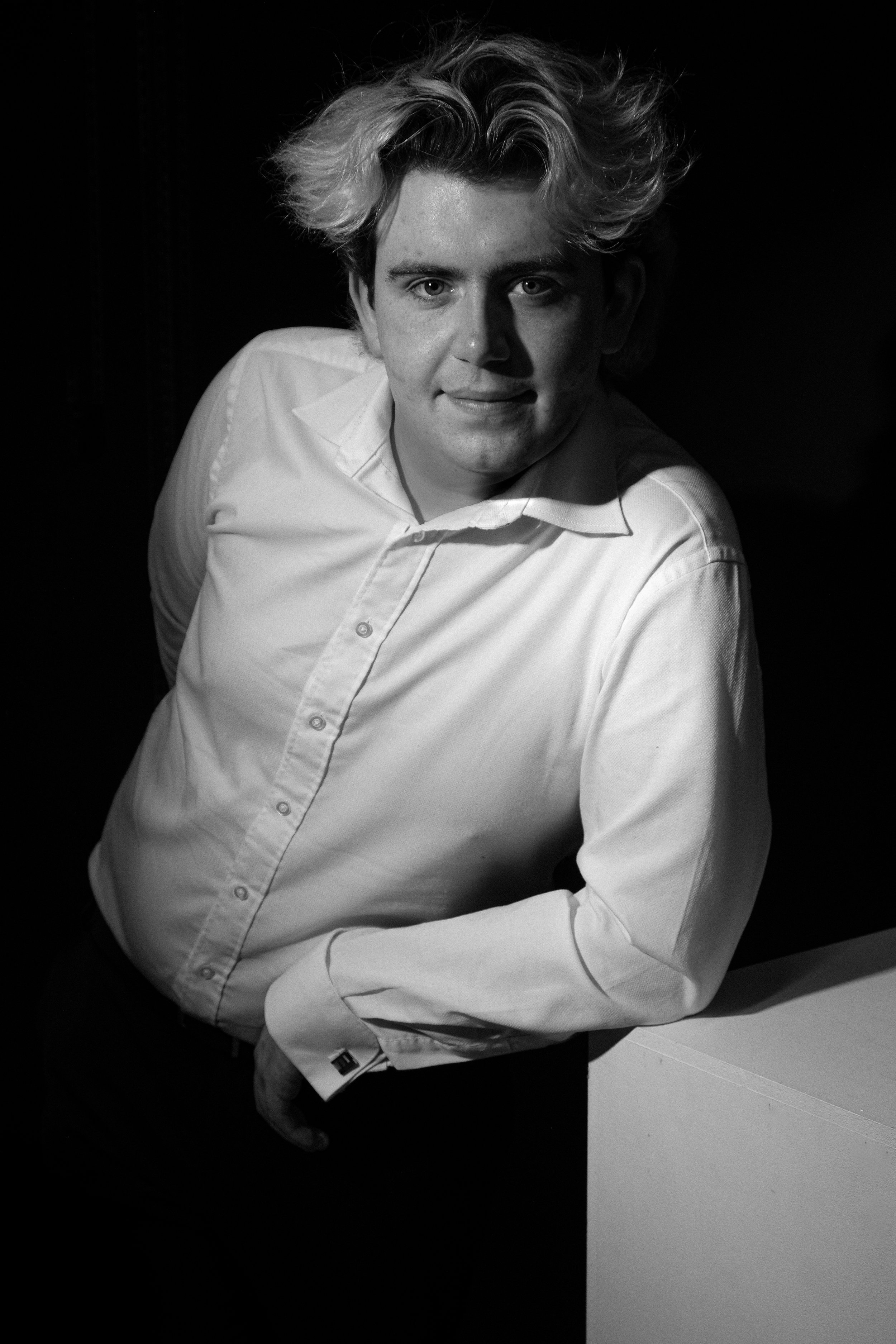 Black and white portrait of a young man with wavy hair in a white shirt, leaning on a table with a dark background.