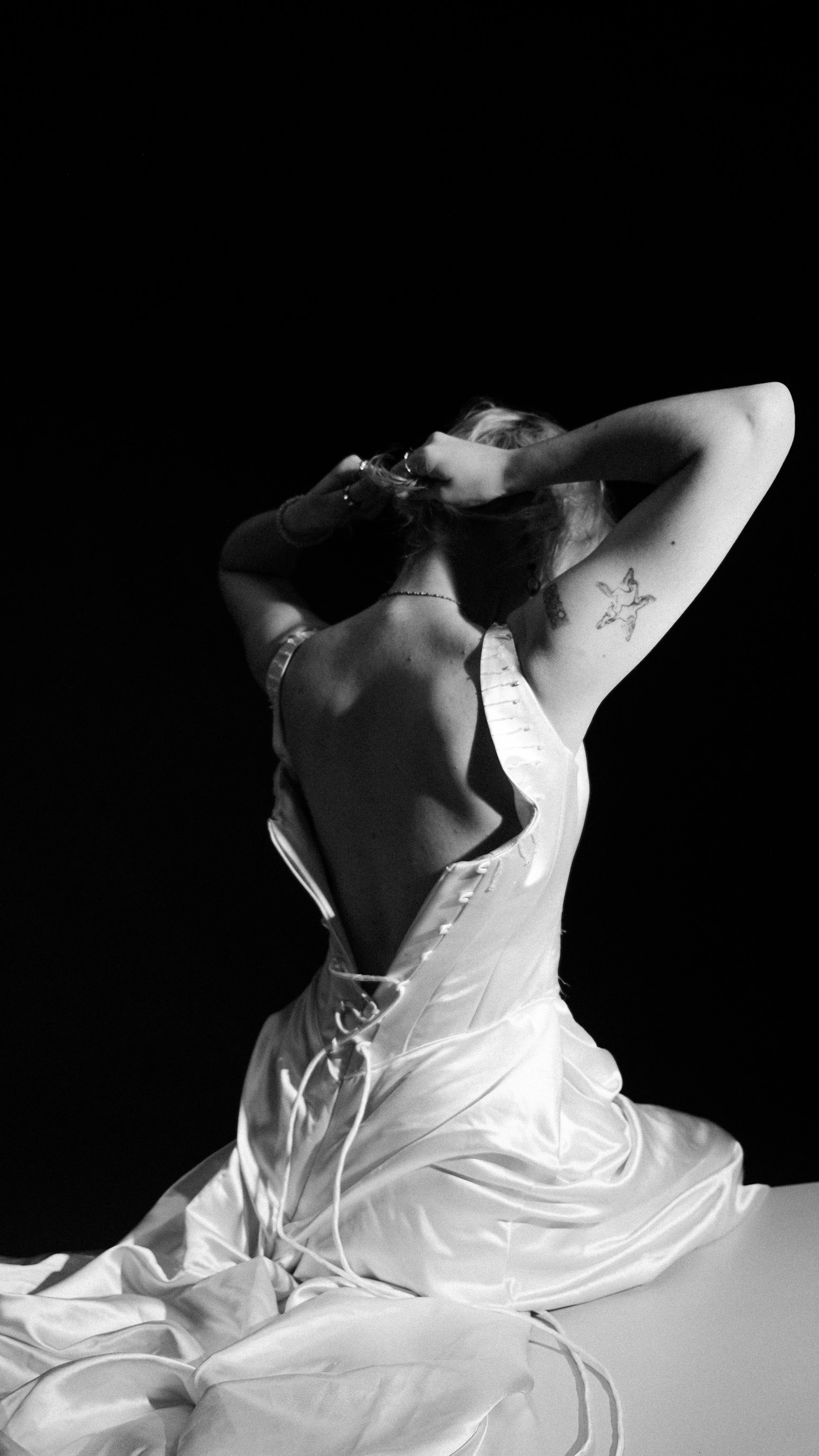 Black and white photo of a woman sitting on the floor, back slightly arched, with her hands in her hair. She is wearing a satin dress with an open back and has a tattoo of a star on her upper arm.