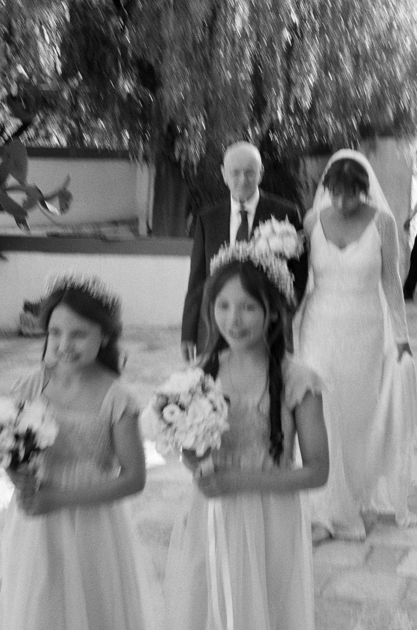 Black and white photo of a wedding scene showing a group of people, including a man in a suit with a boutonniere, a woman in a wedding gown with a veil, and two young girls in dresses holding bouquets.