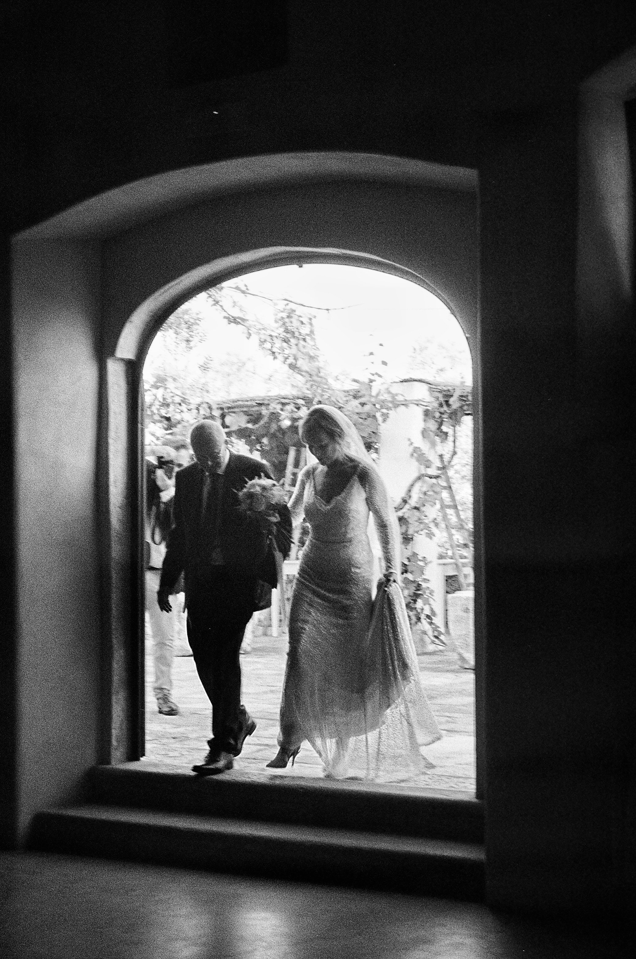 Black and white photo of a bride and an older man, possibly her father, walking through an arched doorway outside during a wedding.
