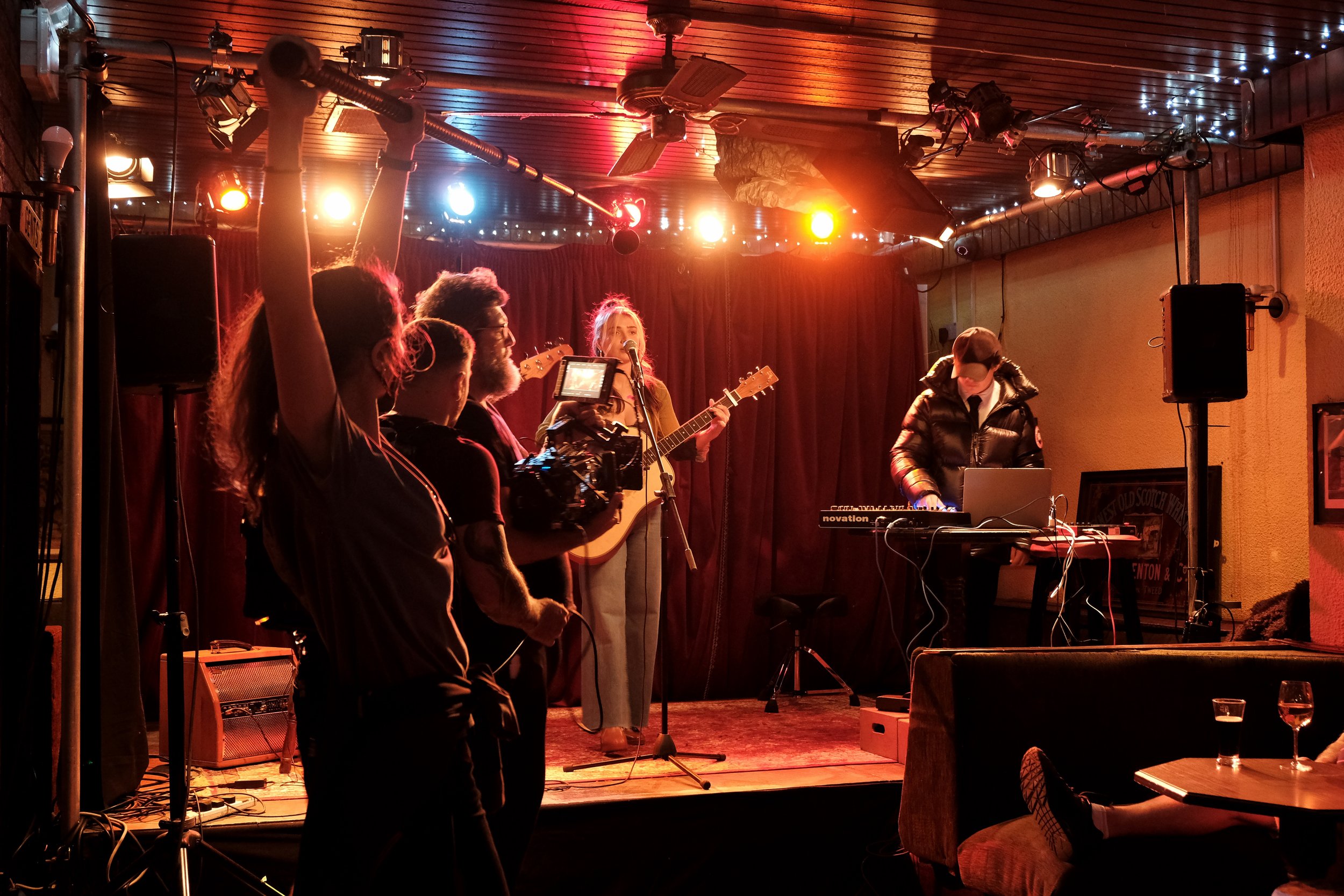 A band performing live on a small stage in a cozy venue, with a woman playing guitar and singing into a microphone, a DJ at a table with electronic equipment, and a camera crew filming the performance. The background has red velvet curtains and color