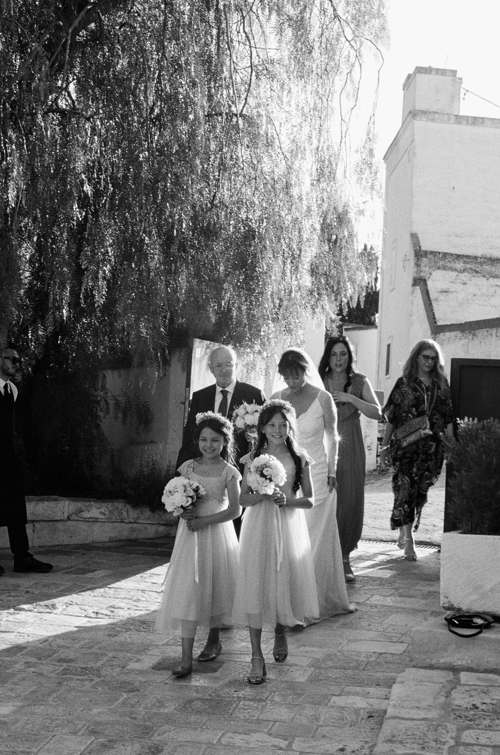 Black and white photo of a wedding procession with two young girls in white dresses holding bouquets, followed by adults, outdoors on a sunny day.