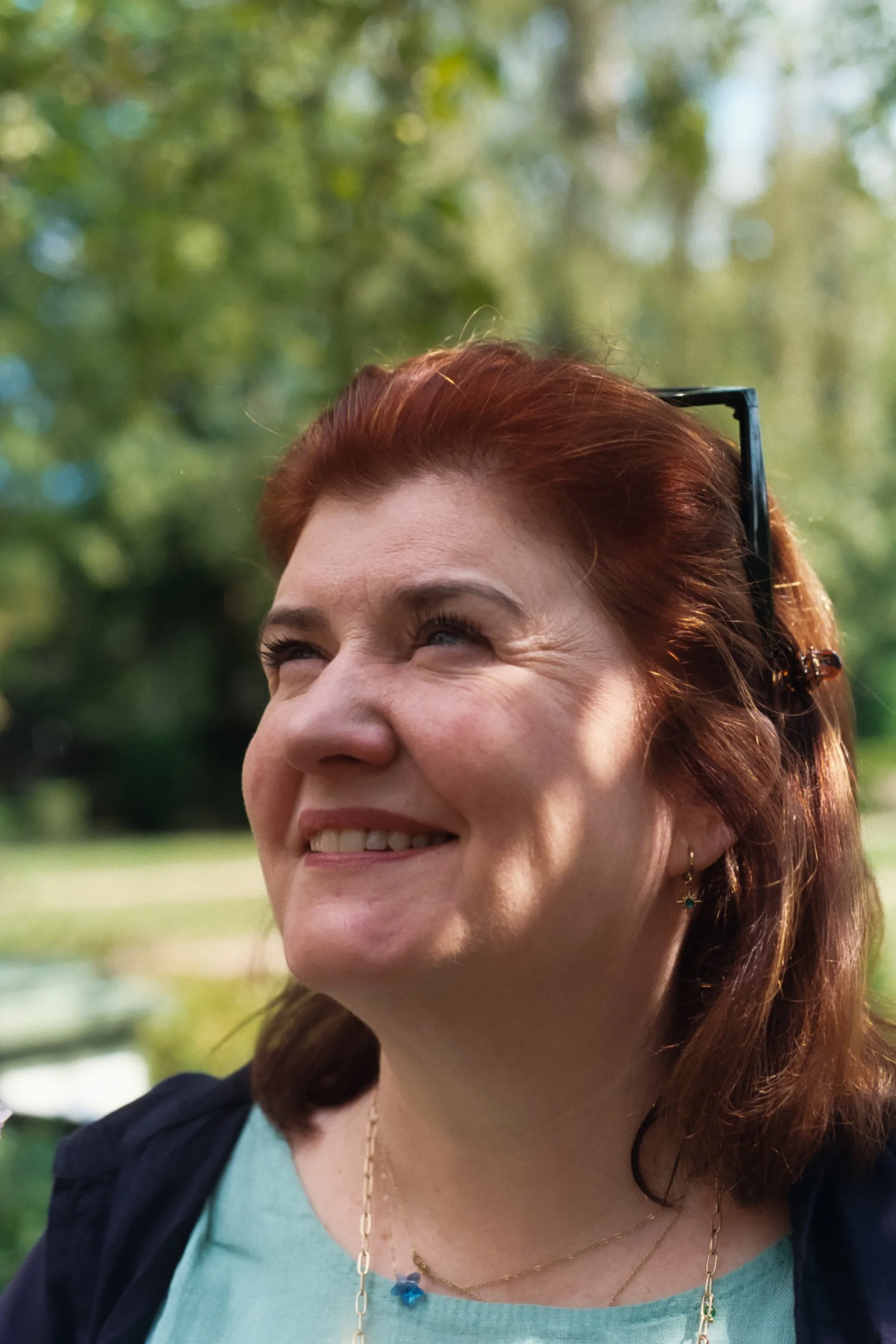 A woman with shoulder-length red hair and sunglasses on her head, smiling outdoors with a background of green trees and sunlight.