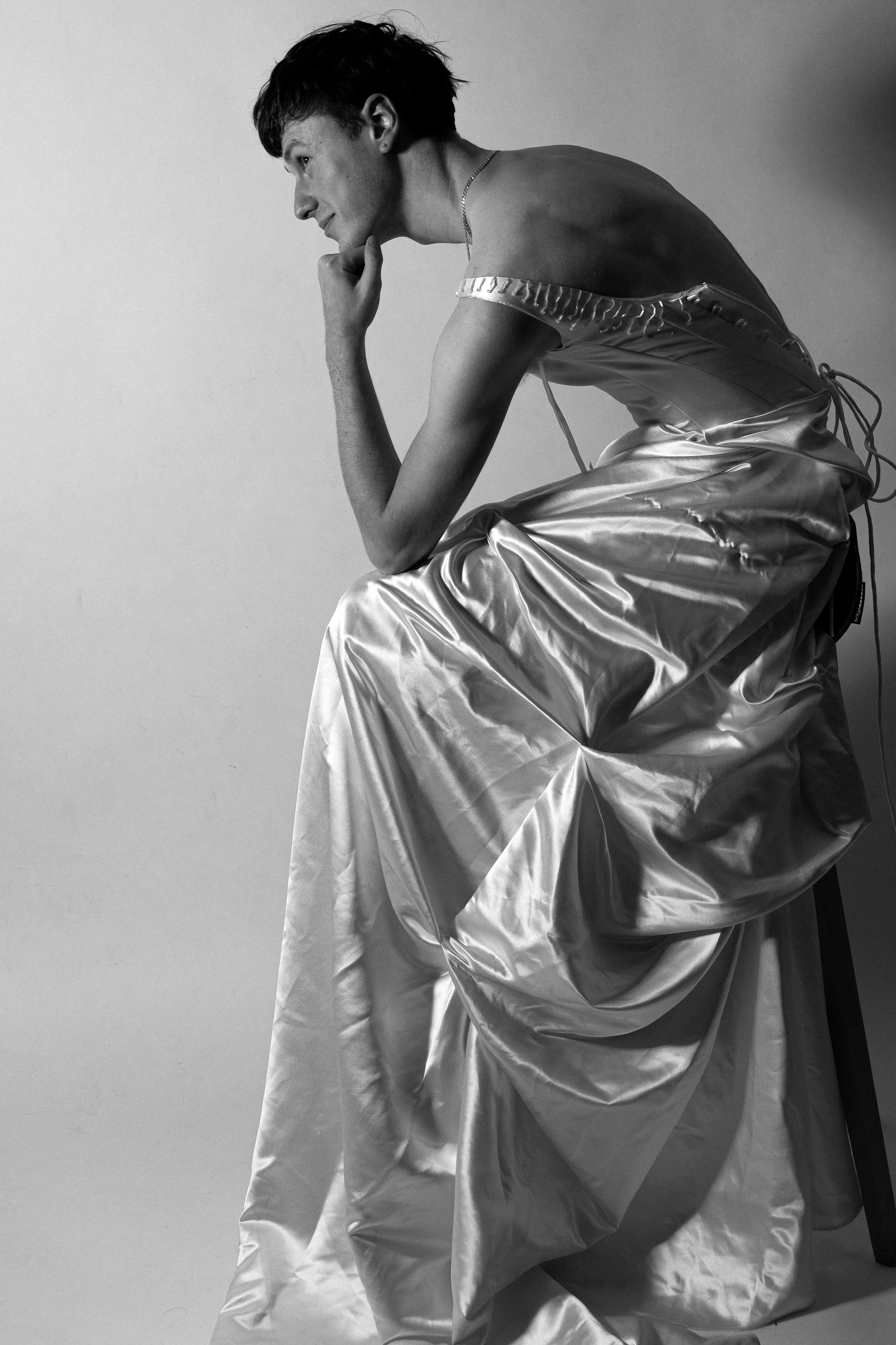 A person in a satin gown sitting on a stool, resting their chin on their hand in a reflective pose, shot in black and white.