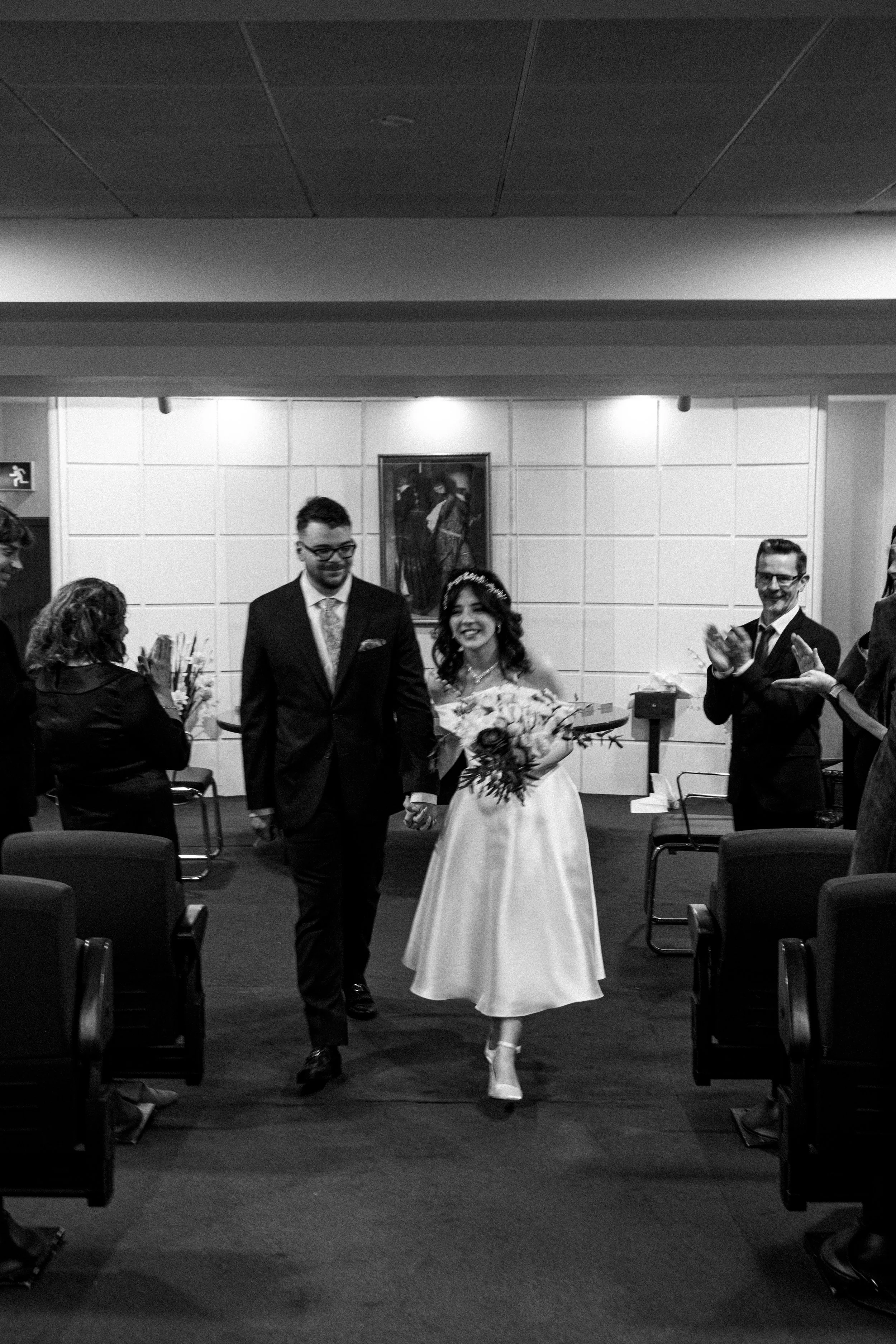 Black and white photograph of a newlywed couple walking down the aisle, holding hands, with smiling guests clapping and congratulating them.