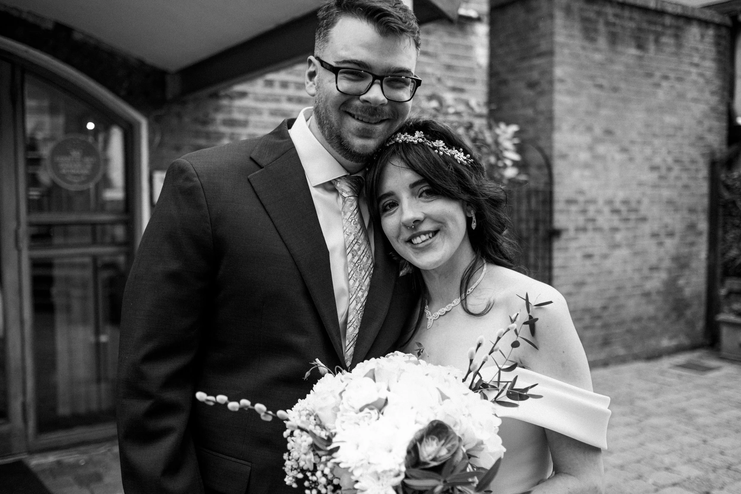 Black and white photo of a bride and groom smiling, with the bride holding a bouquet, standing outside a brick building.