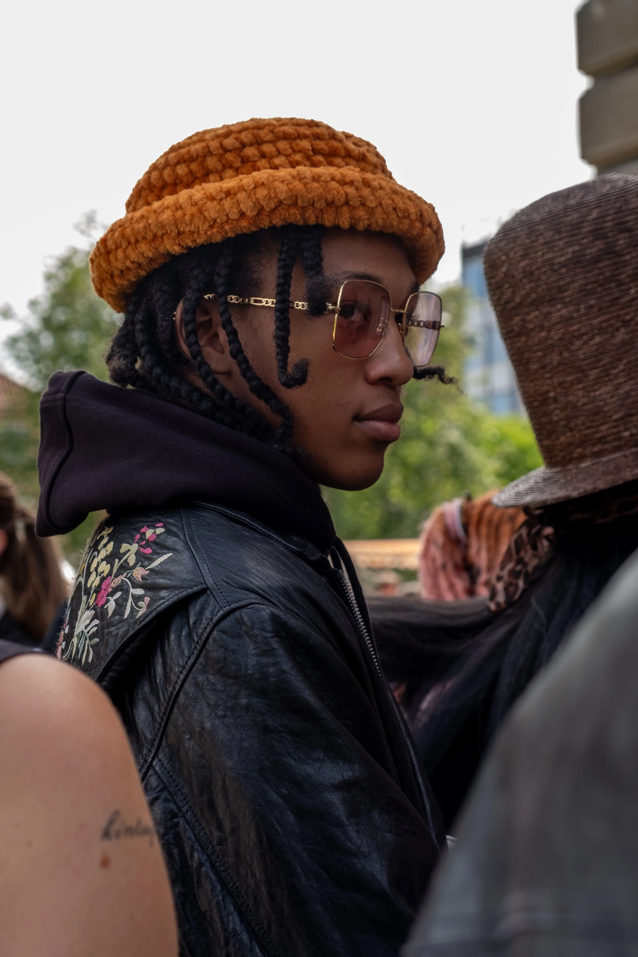 Young man with dark braids wearing sunglasses, a rust-colored knitted hat, a black leather jacket with floral embroidery on the shoulder, and a black hoodie underneath, outdoors with blurred trees and buildings in the background.