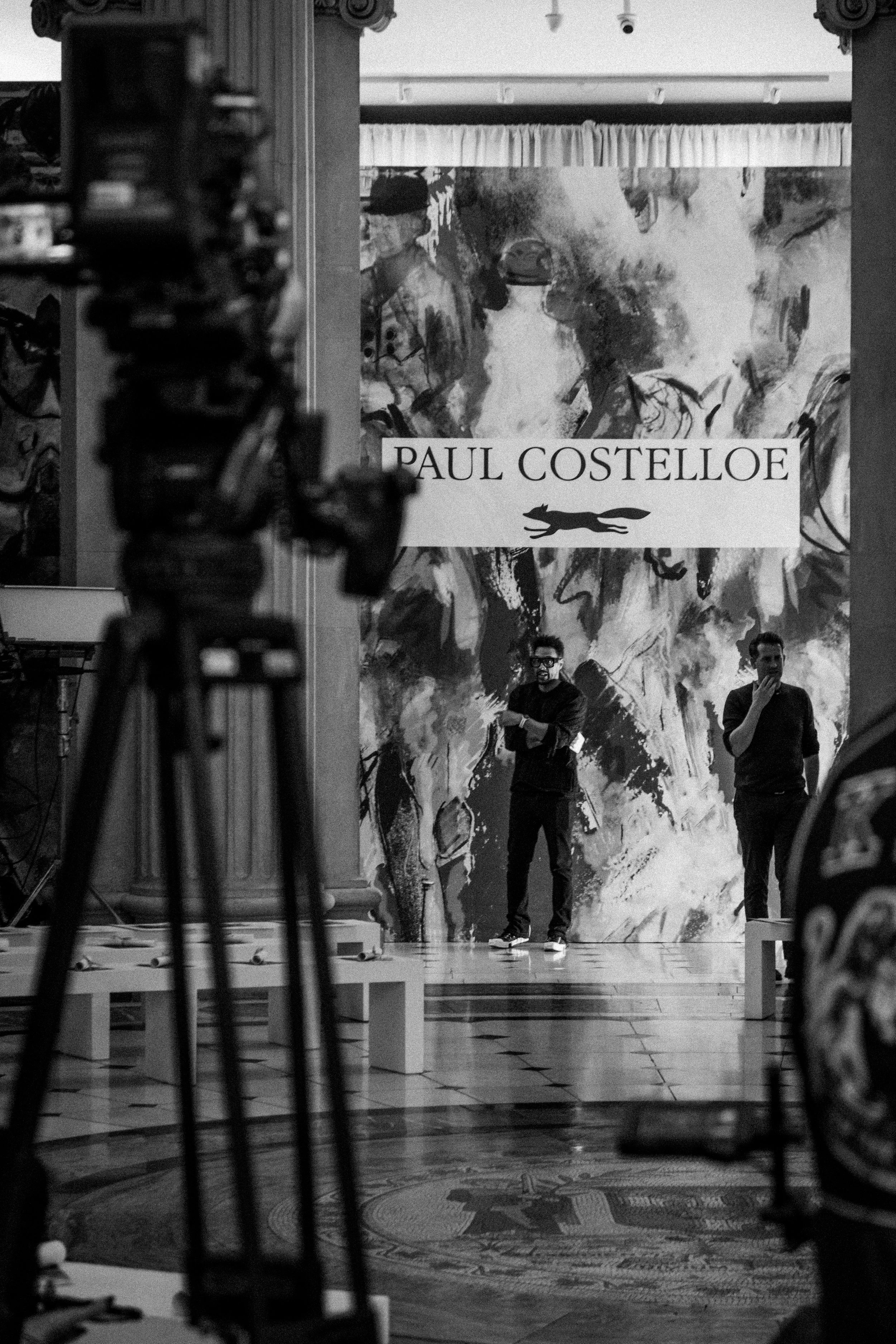 Gallery or art exhibition space with a large abstract painting in the background and a banner reading 'Paul Costelloe.' Two men stand in front of the painting, one speaking into a microphone and the other touching his face, while a camera on a tripod