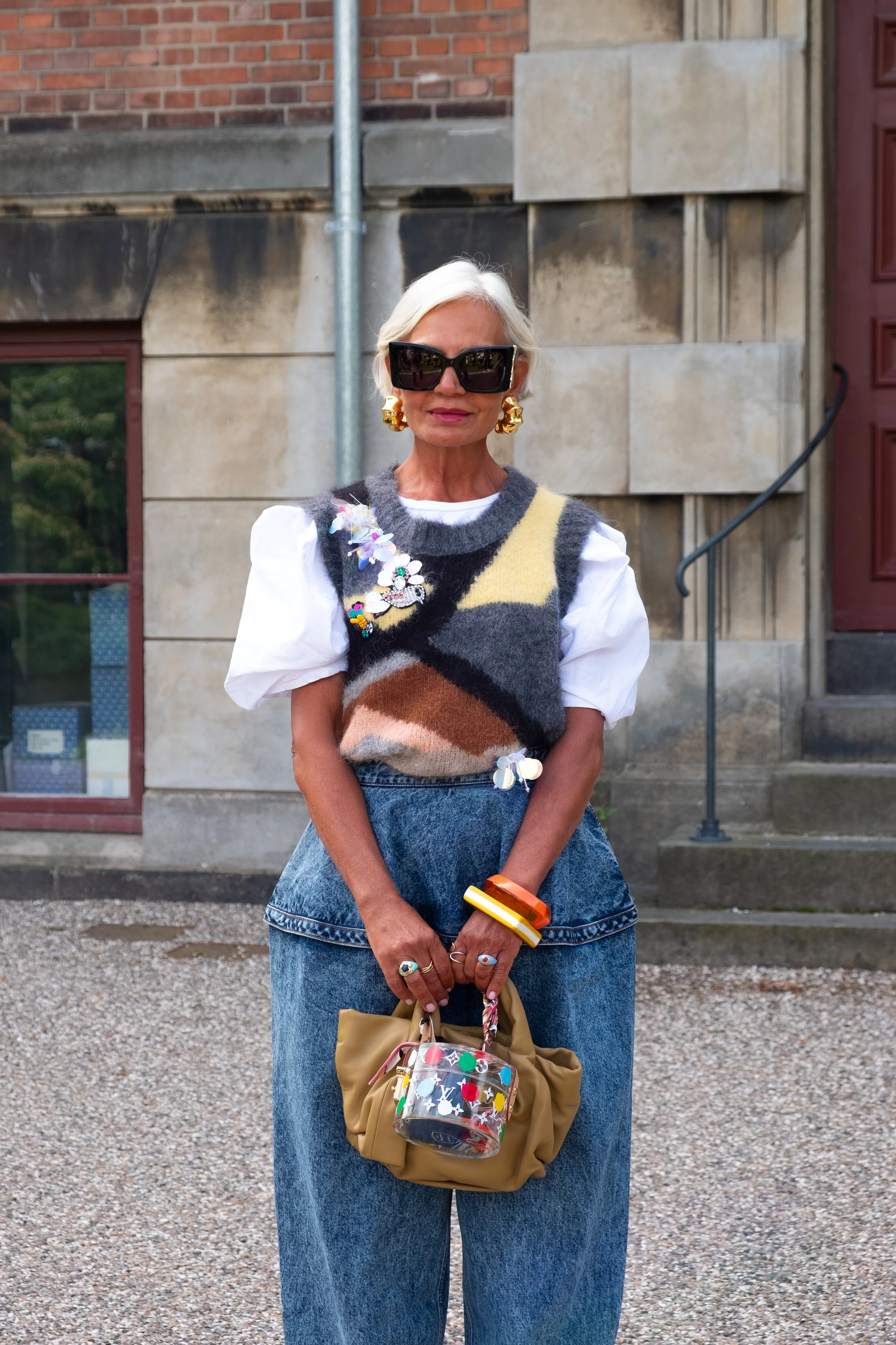 An elderly woman with short white hair wearing large black sunglasses, statement earrings, a multicolored fuzzy sweater vest over a white puff-sleeve blouse, and high-waisted blue jeans, holding a beige bag with a colorful decorative tag, standing in