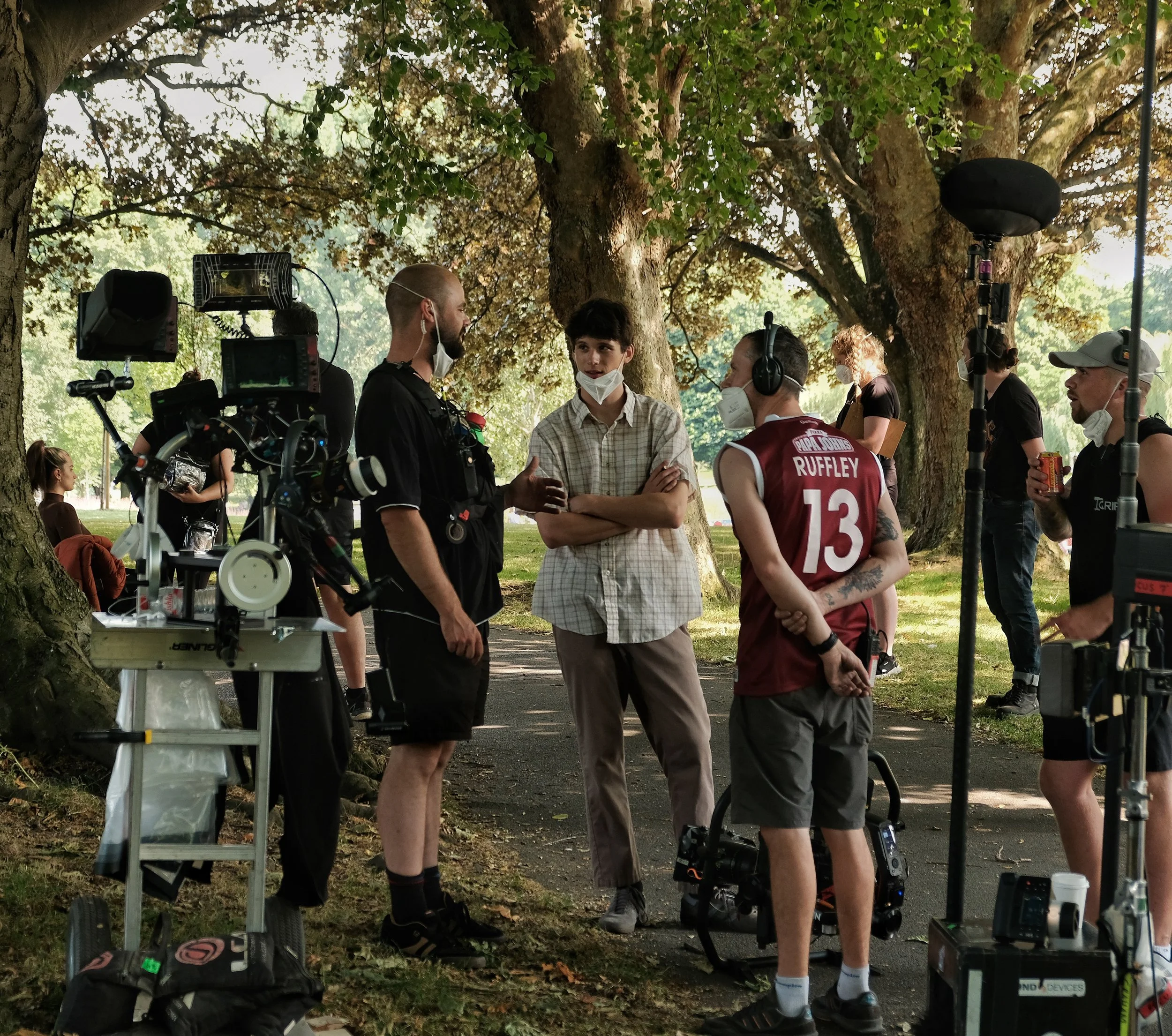 Film crew filming outdoors in park under large trees, with two actors in conversation, crew members wearing headphones and masks, camera equipment and lights set up around them
