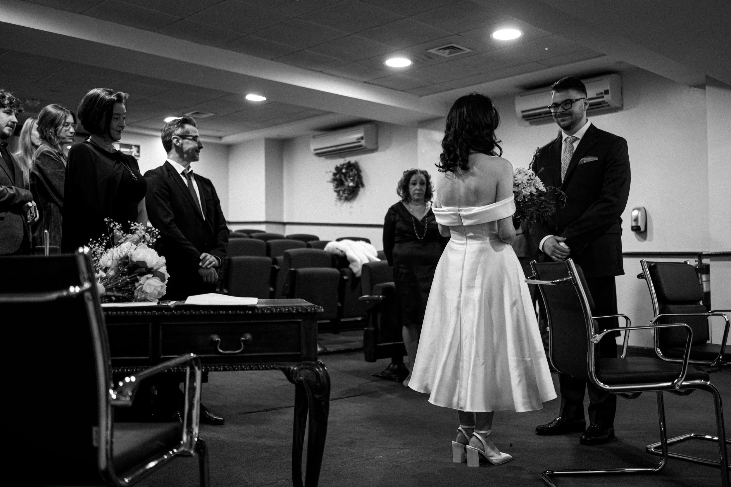 A black and white photo of a wedding ceremony with a bride and groom facing each other, holding flowers, in a room with guests standing and watching.