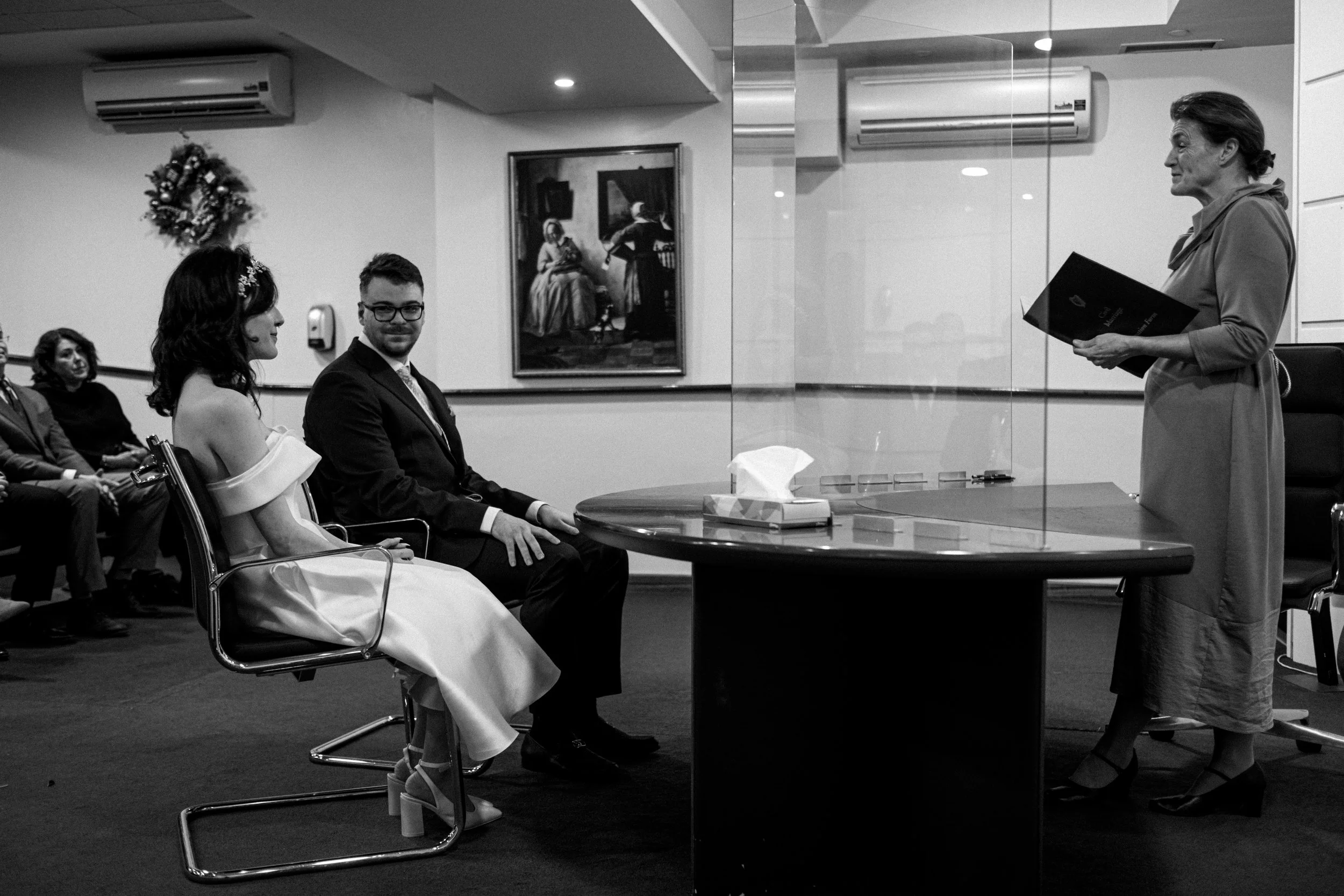 A couple, dressed in wedding attire, sitting in front of an officiant during a wedding ceremony in a decorated indoor venue, with guests seated in the background.