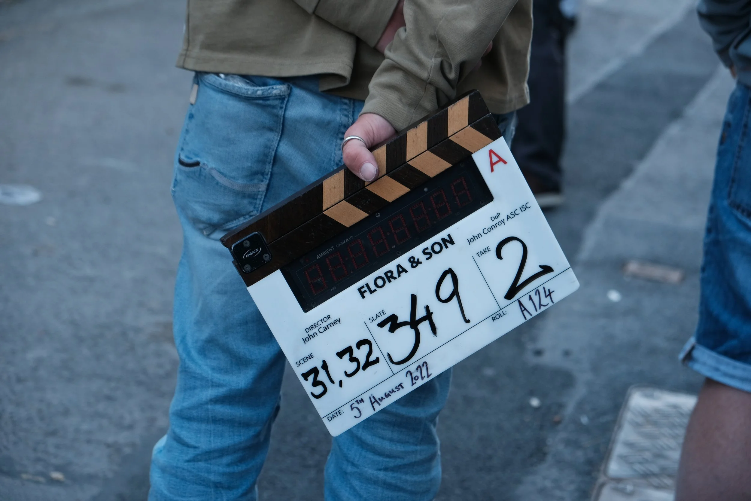 Person holding a film slate with the title 'Flora & Son,' scene number 31,32, 349, 2, dated August 5th, 2022, on a street.