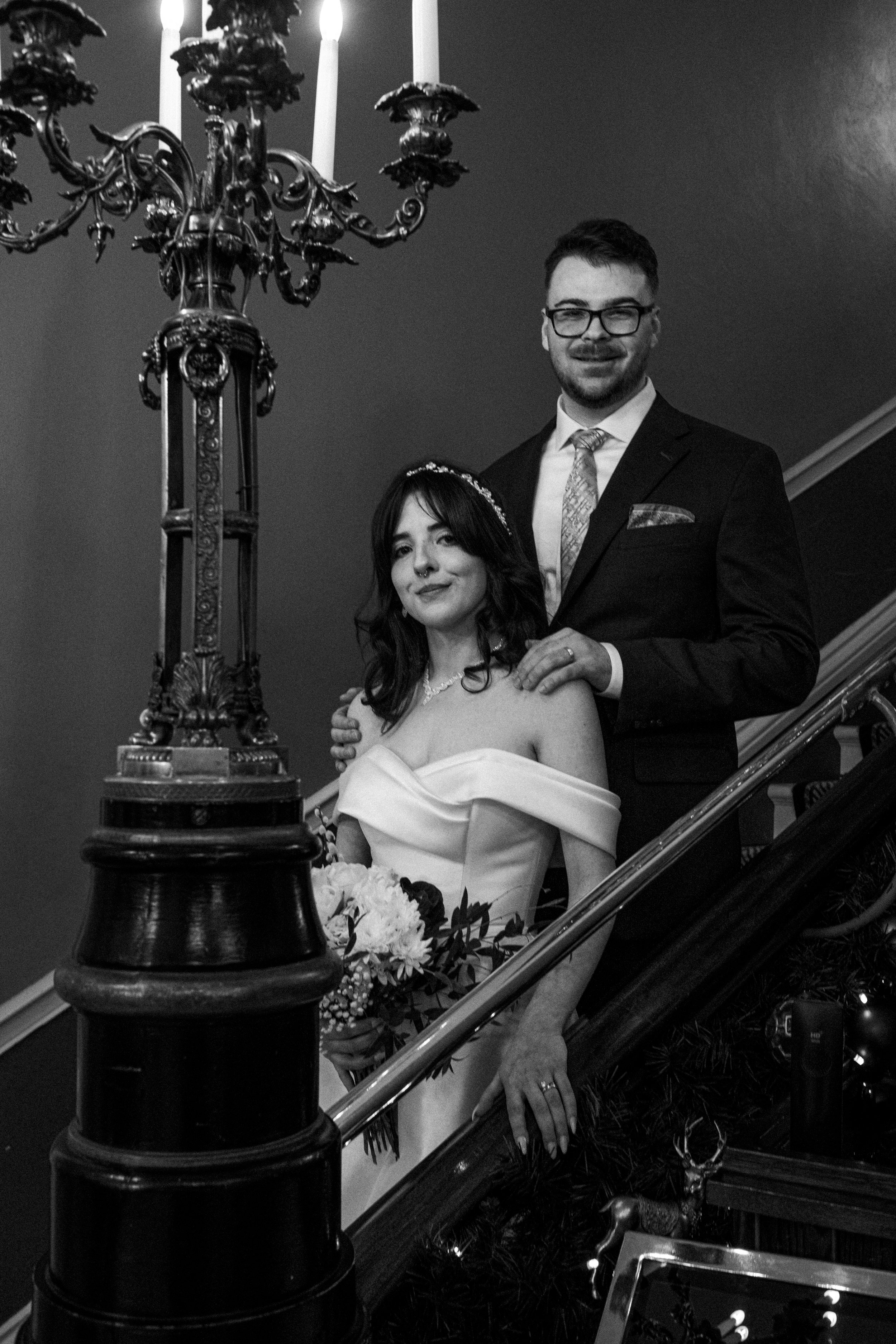 A black and white photo of a man and woman dressed in wedding attire on a staircase. The woman is holding a bouquet and wearing an off-shoulder wedding dress with a flower crown. The man is wearing a suit with a patterned tie and pocket square. They 