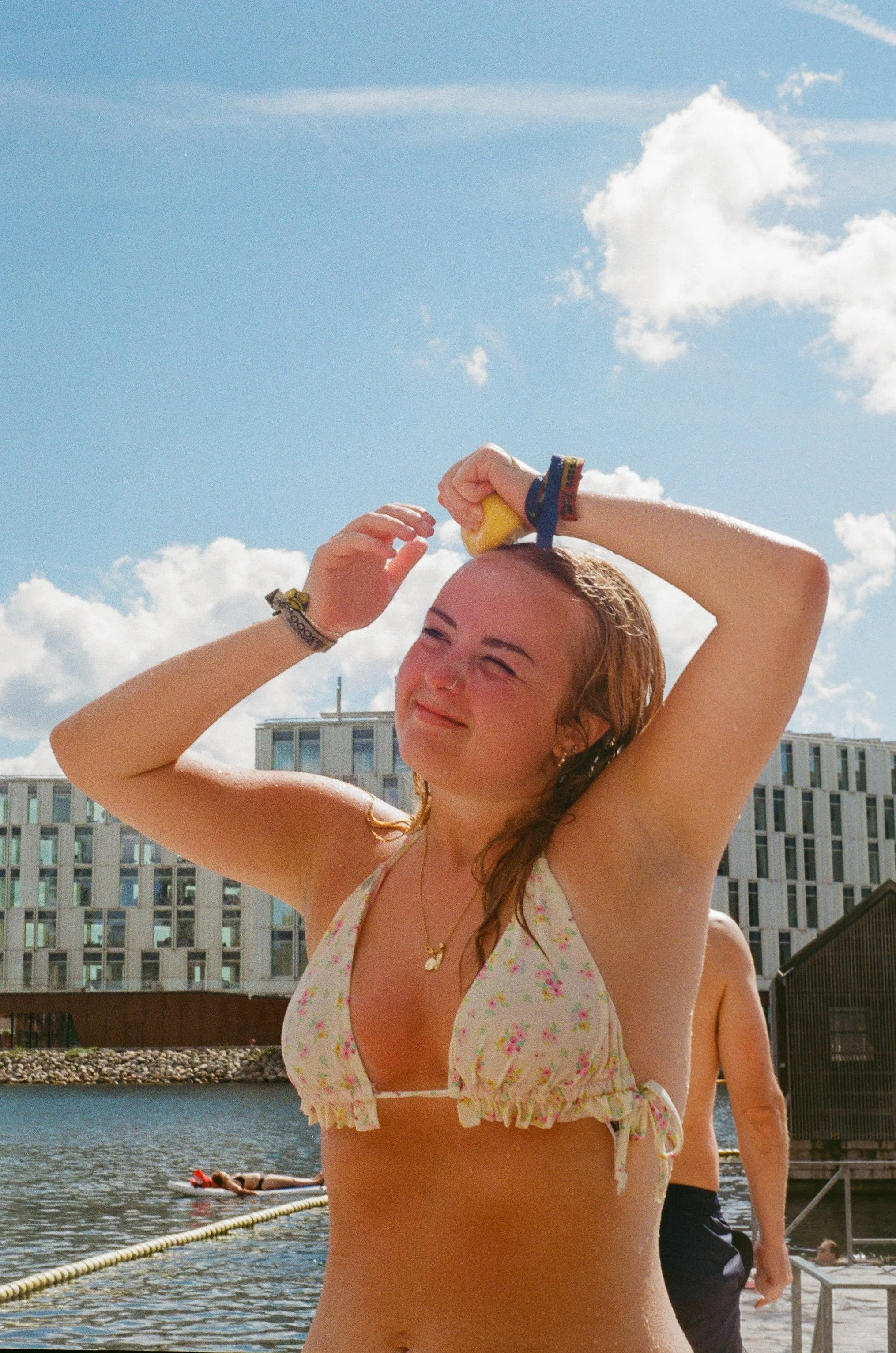 A young woman in a floral bikini top adjusts her wet hair with a yellow sponge while standing outdoors near water, under a blue sky with clouds.