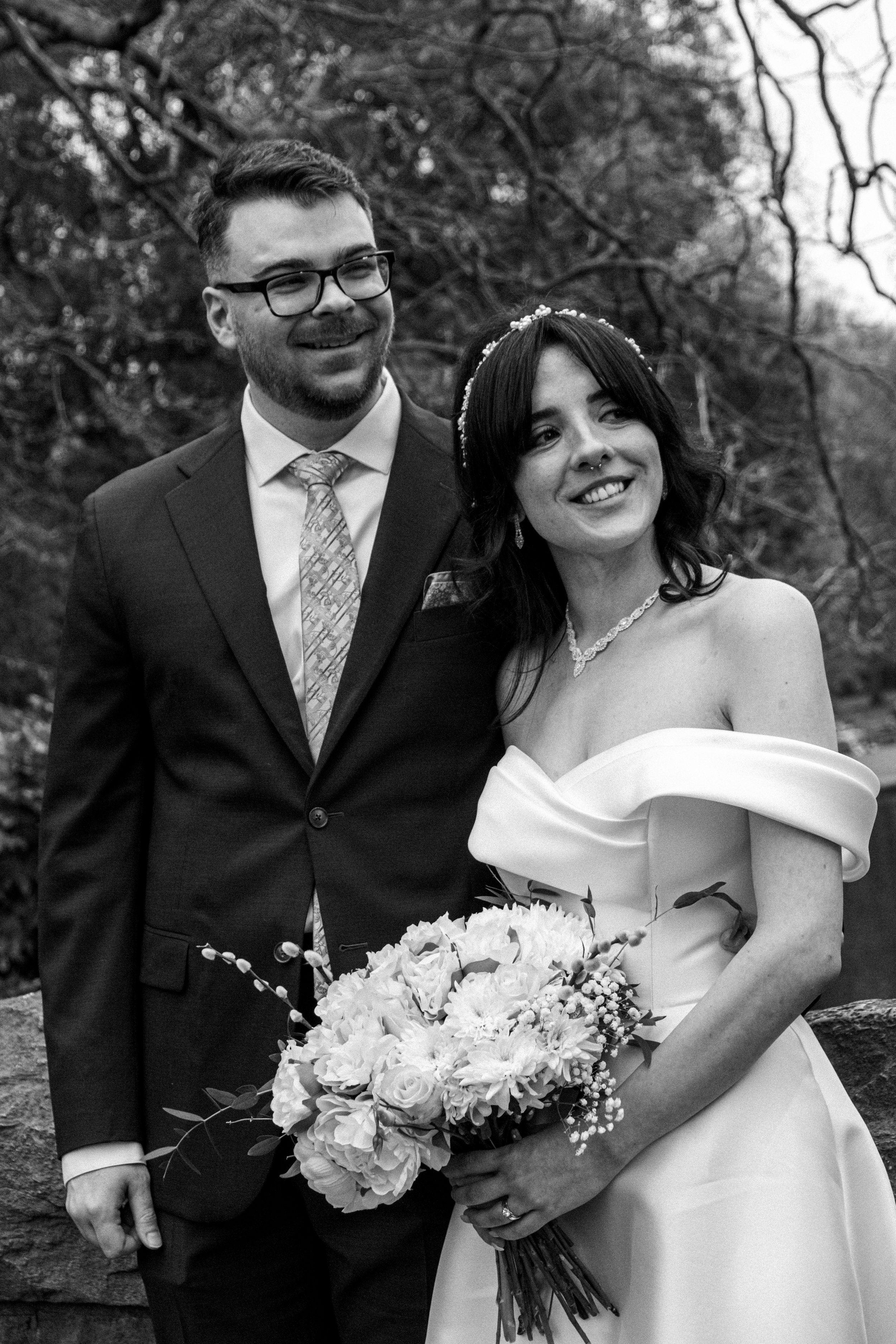 Black and white photo of a bride and groom outdoors, smiling, with trees in the background. The bride is holding a large bouquet of flowers, wearing an off-the-shoulder wedding gown with jewelry, and the groom is dressed in a suit with a tie and glas