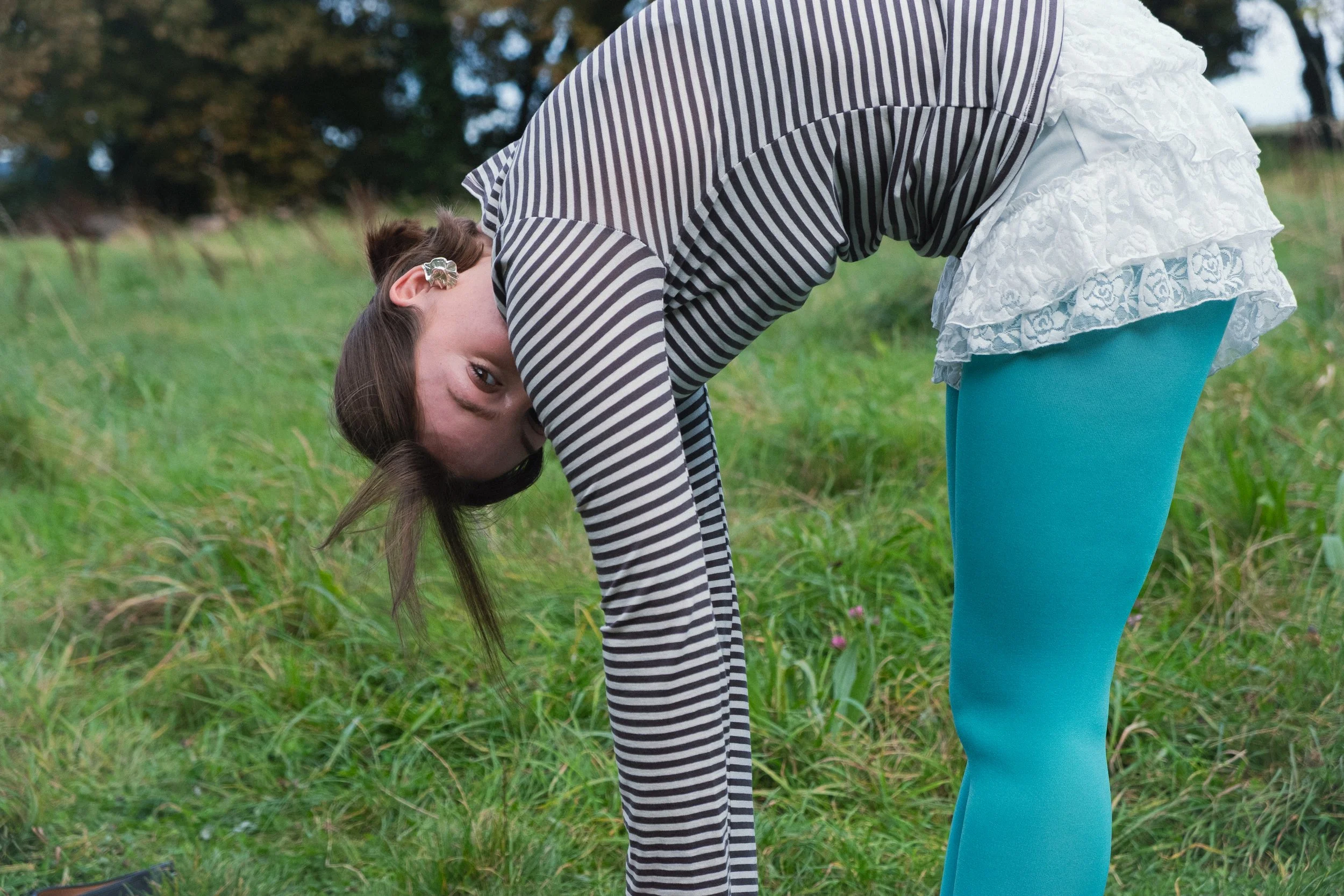 A girl with long brown hair, wearing a striped long-sleeve shirt, white ruffled skirt, and turquoise leggings, is outdoors in a grassy area, bent over with her head upside down, looking at the camera.