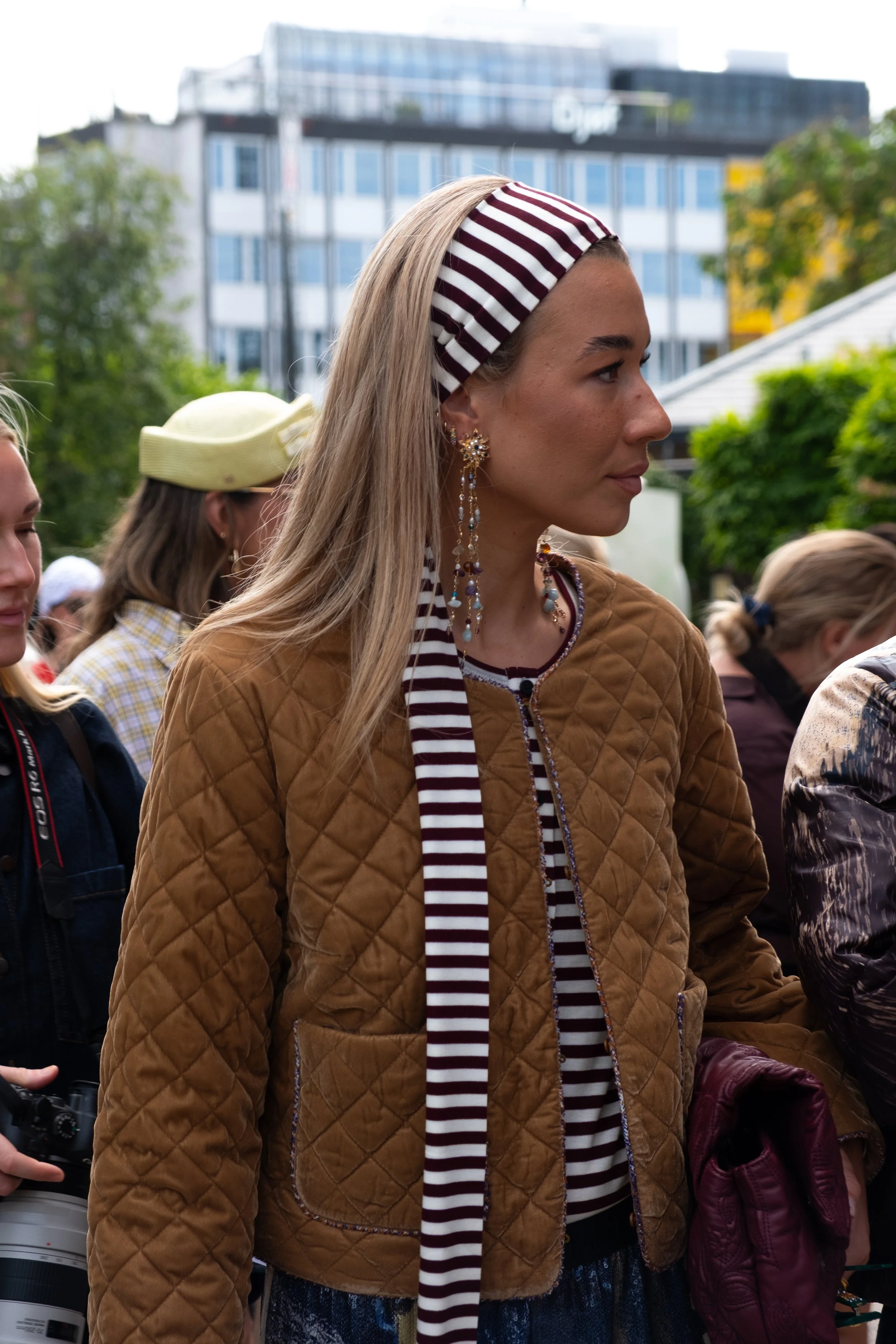 A woman in a brown quilted jacket wearing gold earrings and a striped headscarf at an outdoor event, with other people and buildings in the background.