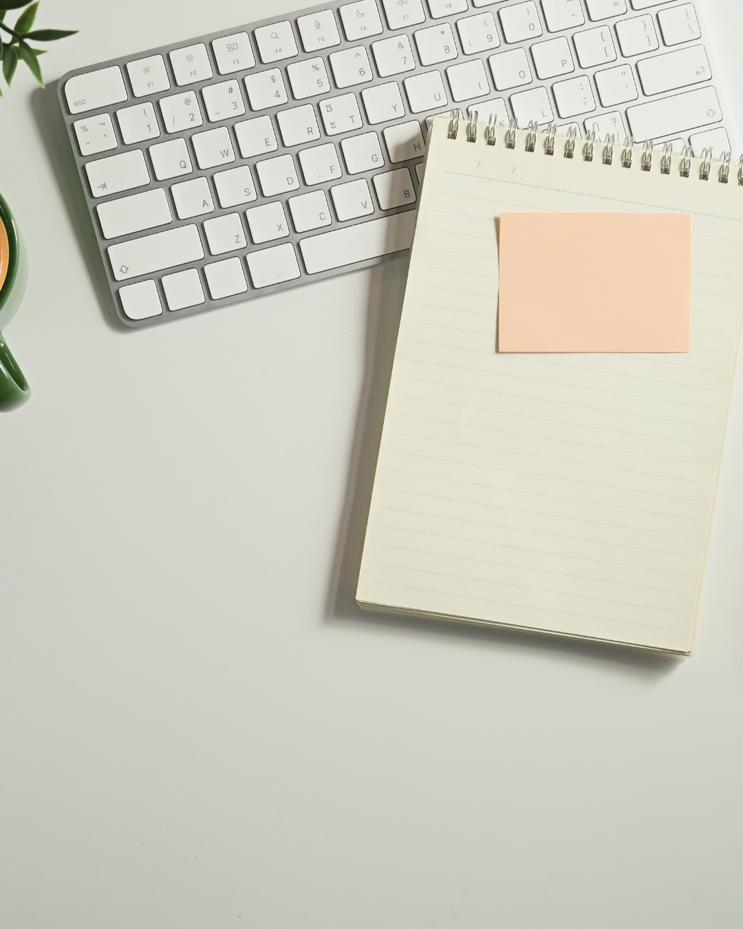 White computer keyboard, spiral notepad with a pink sticky note on top, and a cup with a hot beverage on a white desk.
