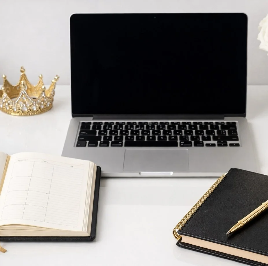 A desk setup with a silver laptop, an open planner, a closed black notebook with a gold pen, a gold crown decoration, and a white flower on a white surface.